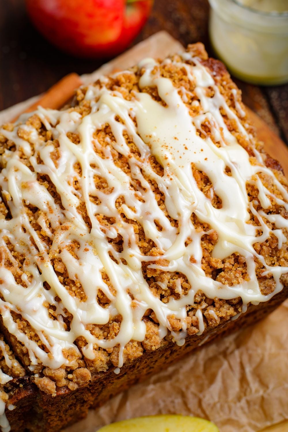 Apple Cinnamon Loaf Cake on parchment paper on a wooden table with a glass of glaze to side and red apple