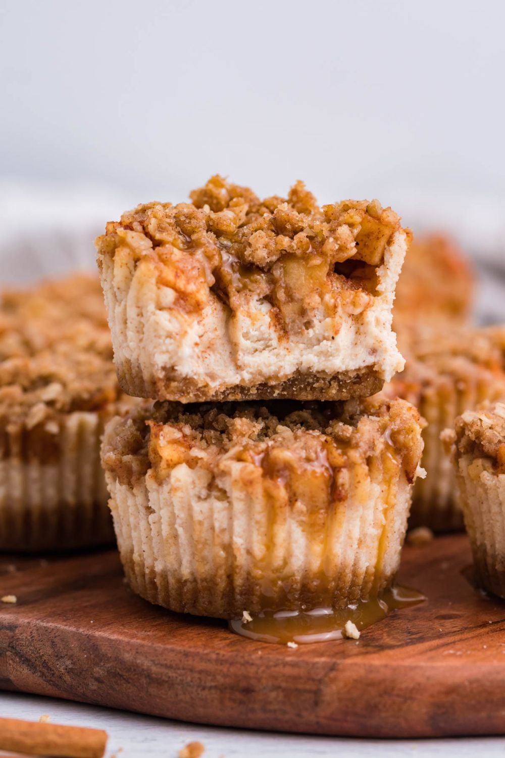 cutting board on marble table with two Apple Crisp Cheesecake Bites stacked on top of one another, and top cheesecake has bite taken out of it. Then around the board are more cheesecakes.
