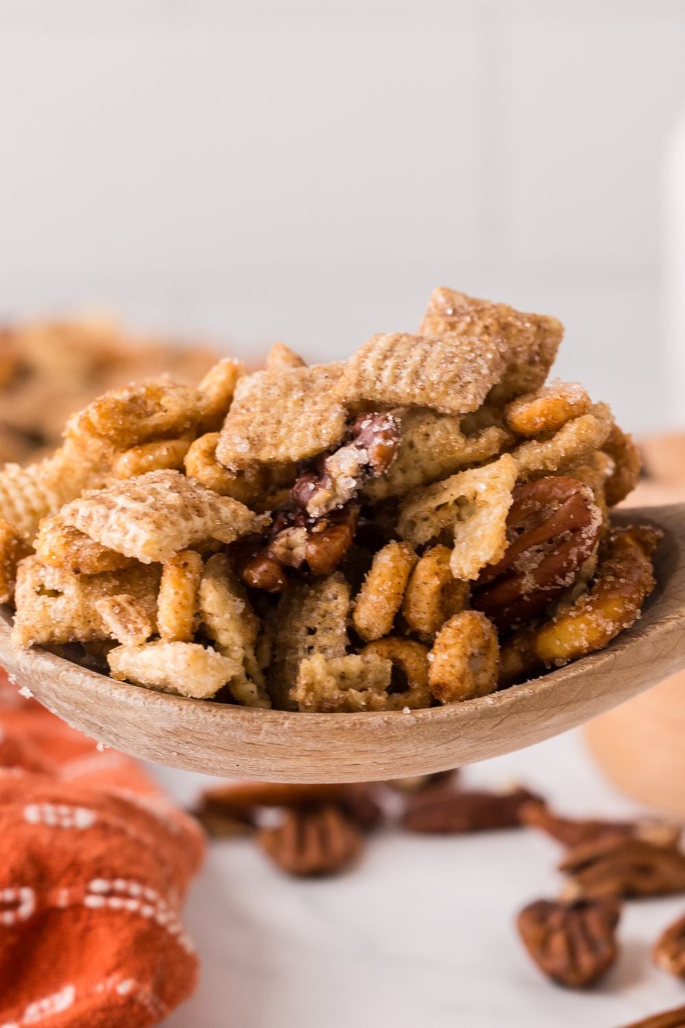 Pumpkin Spice Chex Mix in a wooden spoon with some nuts and more snack mix on the counter with an orange towel in front of table 