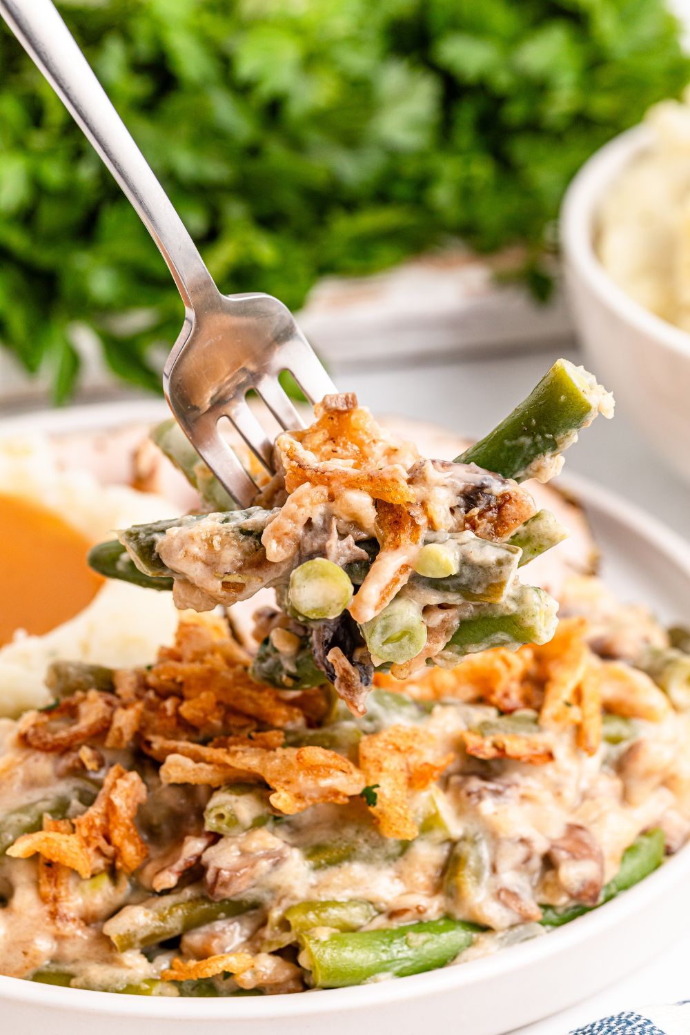 Green Bean Casserole on a white plate on table with a fork scooping up some of the mixture. Parsley greens are behind it on table 
