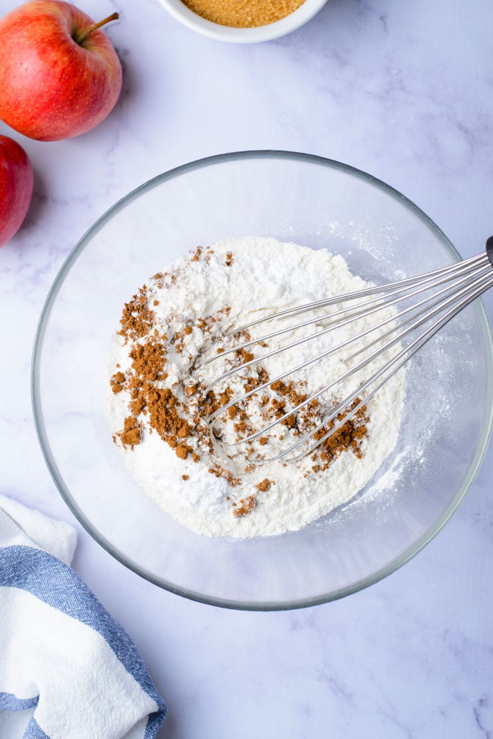 flour and cinnamon mixture in a glass bowl on marble counter with two apples set to the side on table