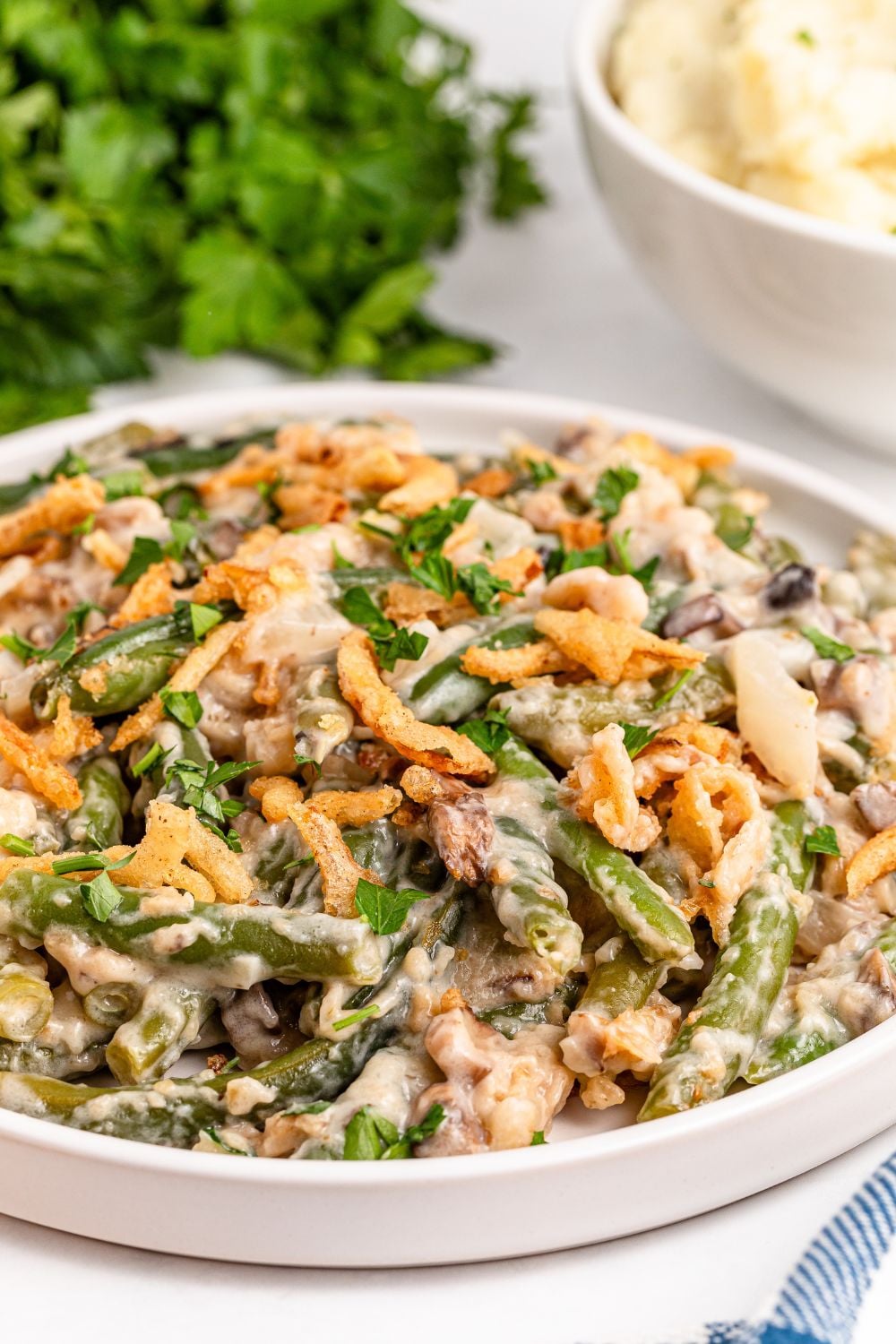 side view of a white plate with an edge sitting on table full of Green Bean Casserole and behind it on table fresh parsley bundle 