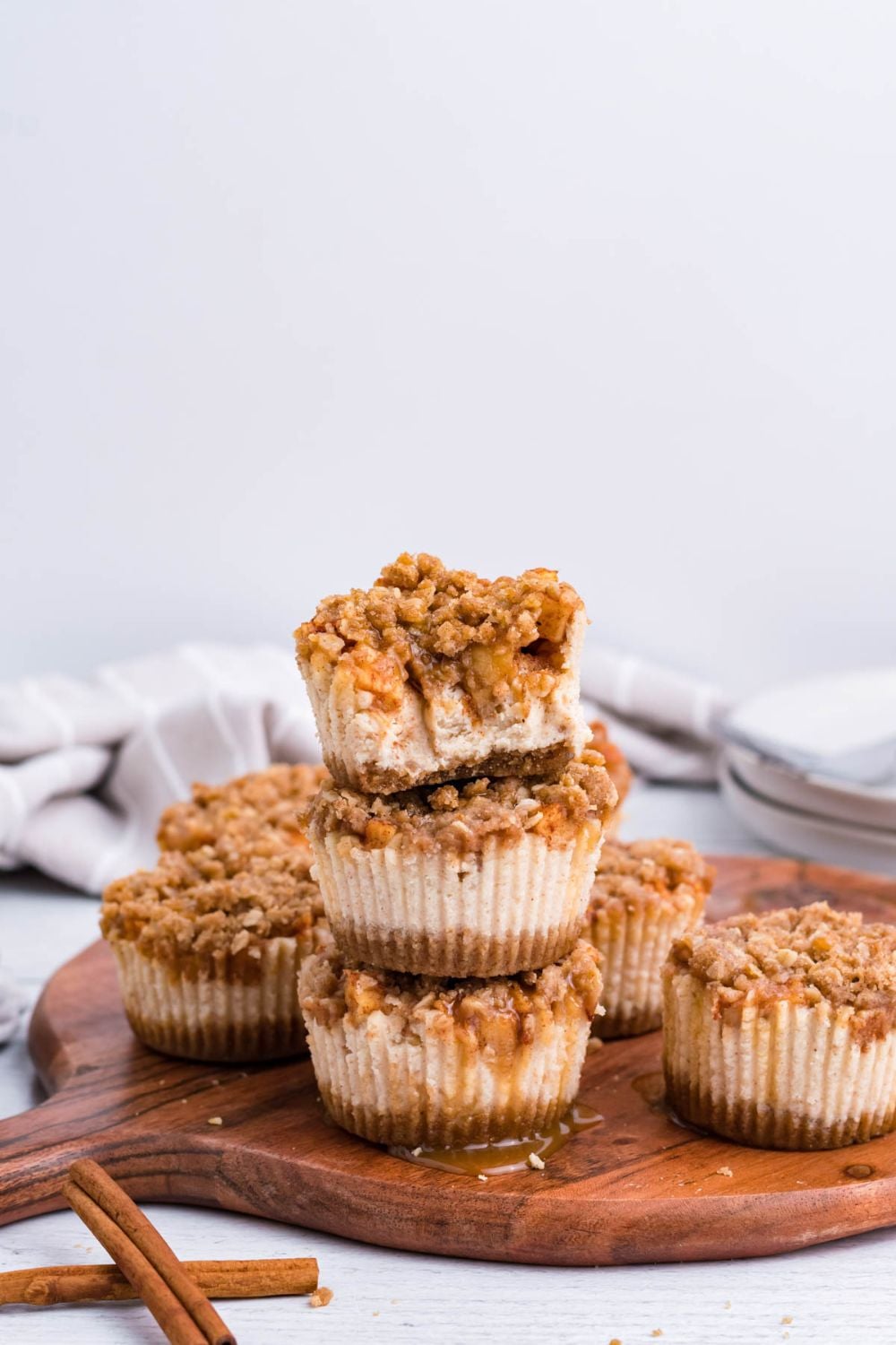 wooden cutting board sitting on marble counter with a towel behind it on table. On the tray is stacks of Apple Crisp Cheesecake Bites with one of the cheesecakes with some bites taken out of it