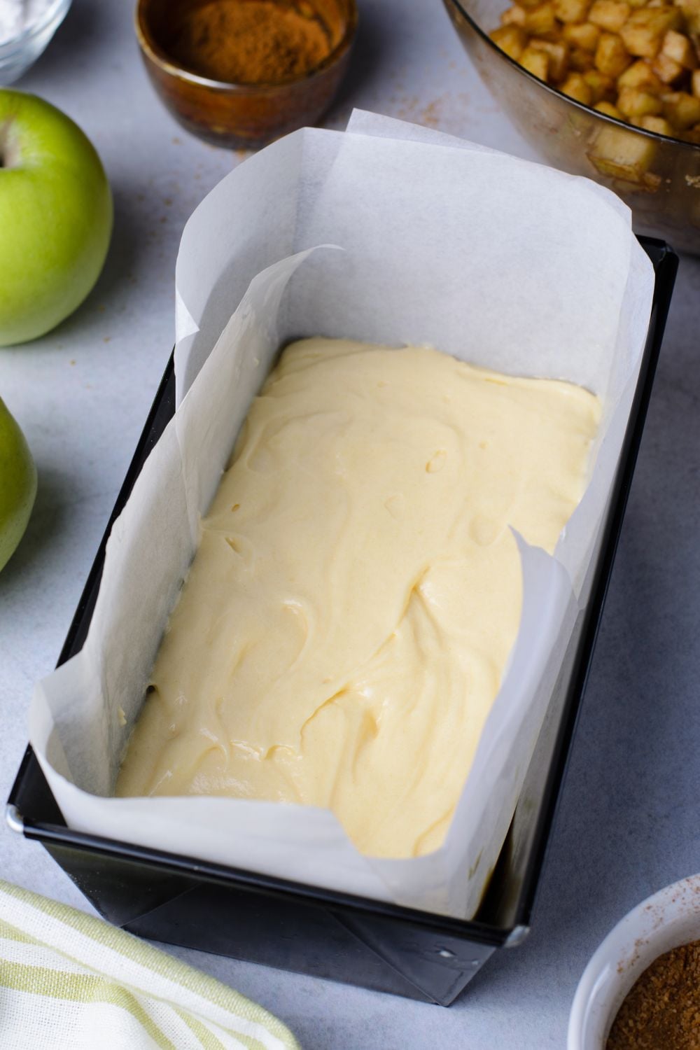 loaf pan with parchment paper in it with a bit of bread batter inside. Apples, nuts, cinnamon and more around the pan to decorate the table