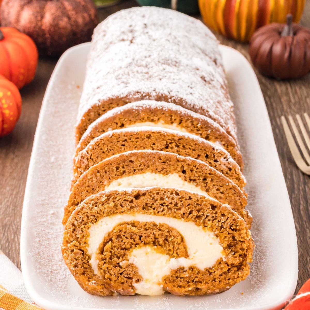 Pumpkin Roll on a long platter sitting on wooden table with pumpkins around to set a fall theme. Part of the pumpkin roll is sliced and the other part is not sliced and dusted with powdered sugar