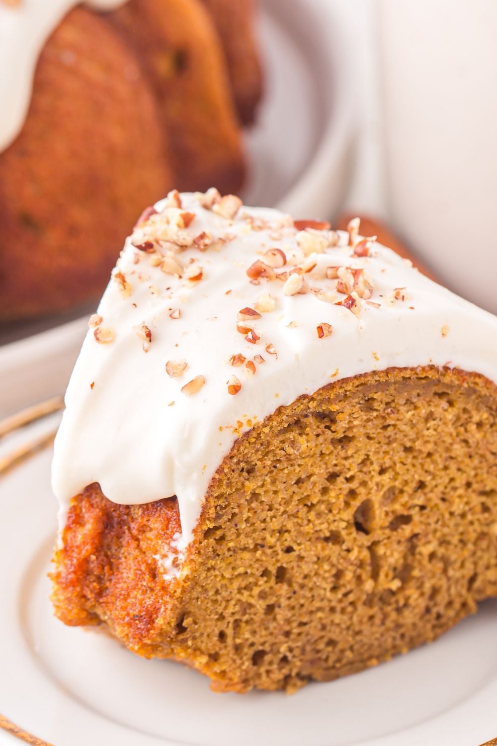 close up view of a slice of Pumpkin Bundt Cake topped with cream cheese glaze and nuts on a white plate sitting on table