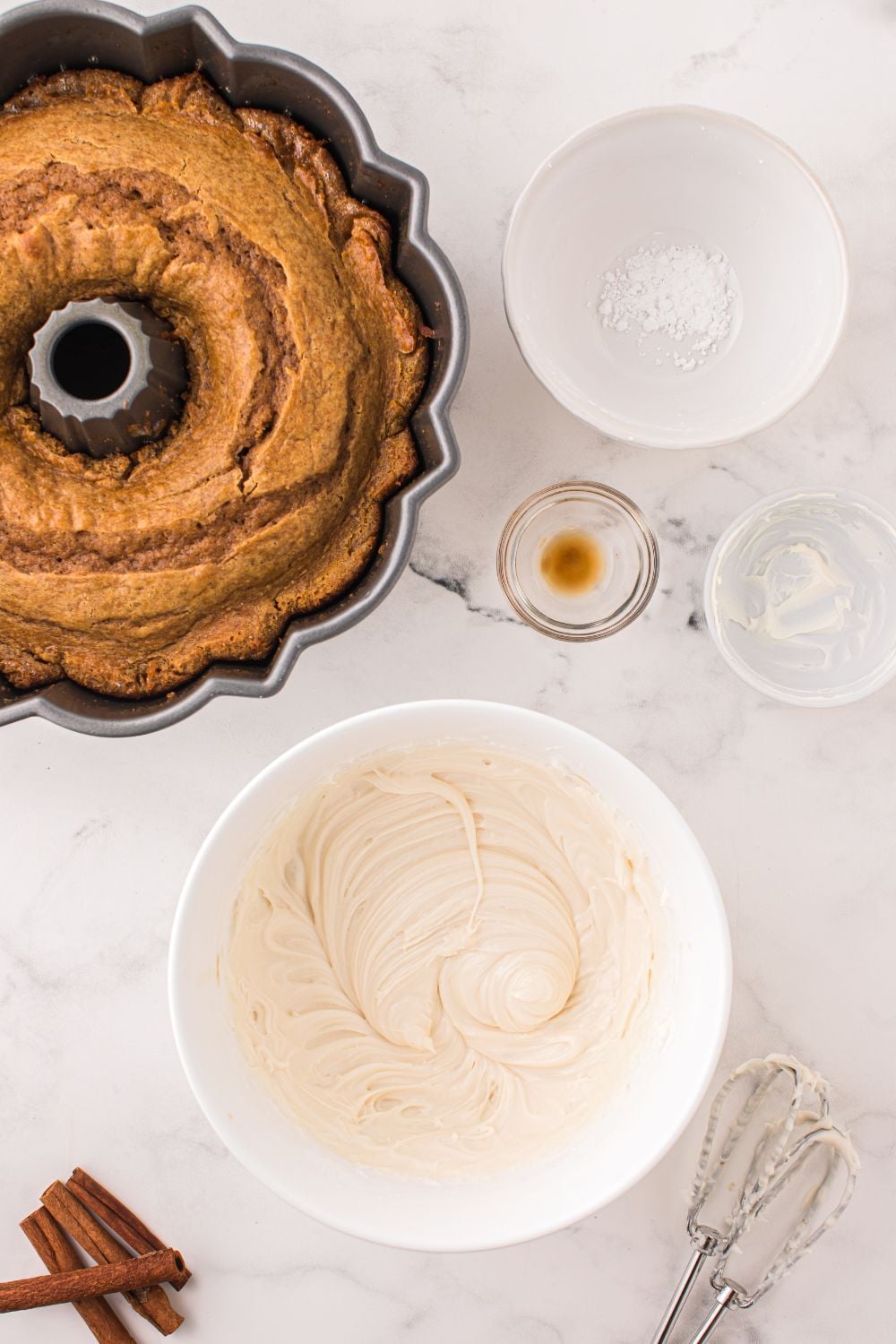 baked pumpkin bundt cake in pan with all the ingredients for cream cheese frosting in bowls around table