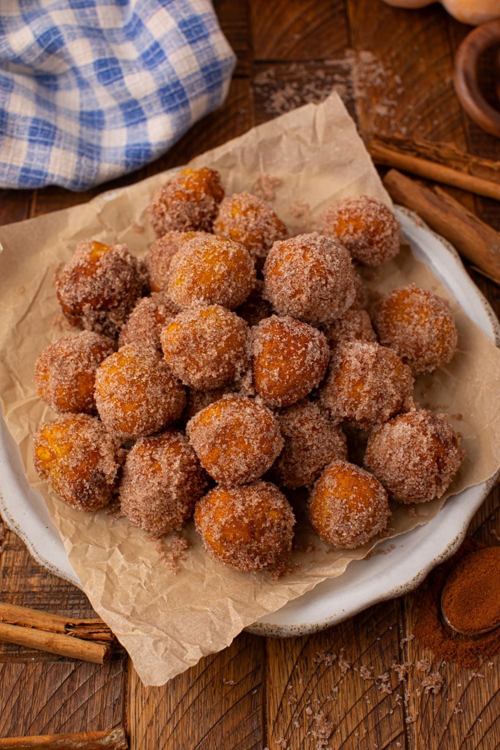 Pumpkin Pie Donut Holes on a platter with a parchment paper lined platter on a wooden table with cinnamon sticks around table and a towel