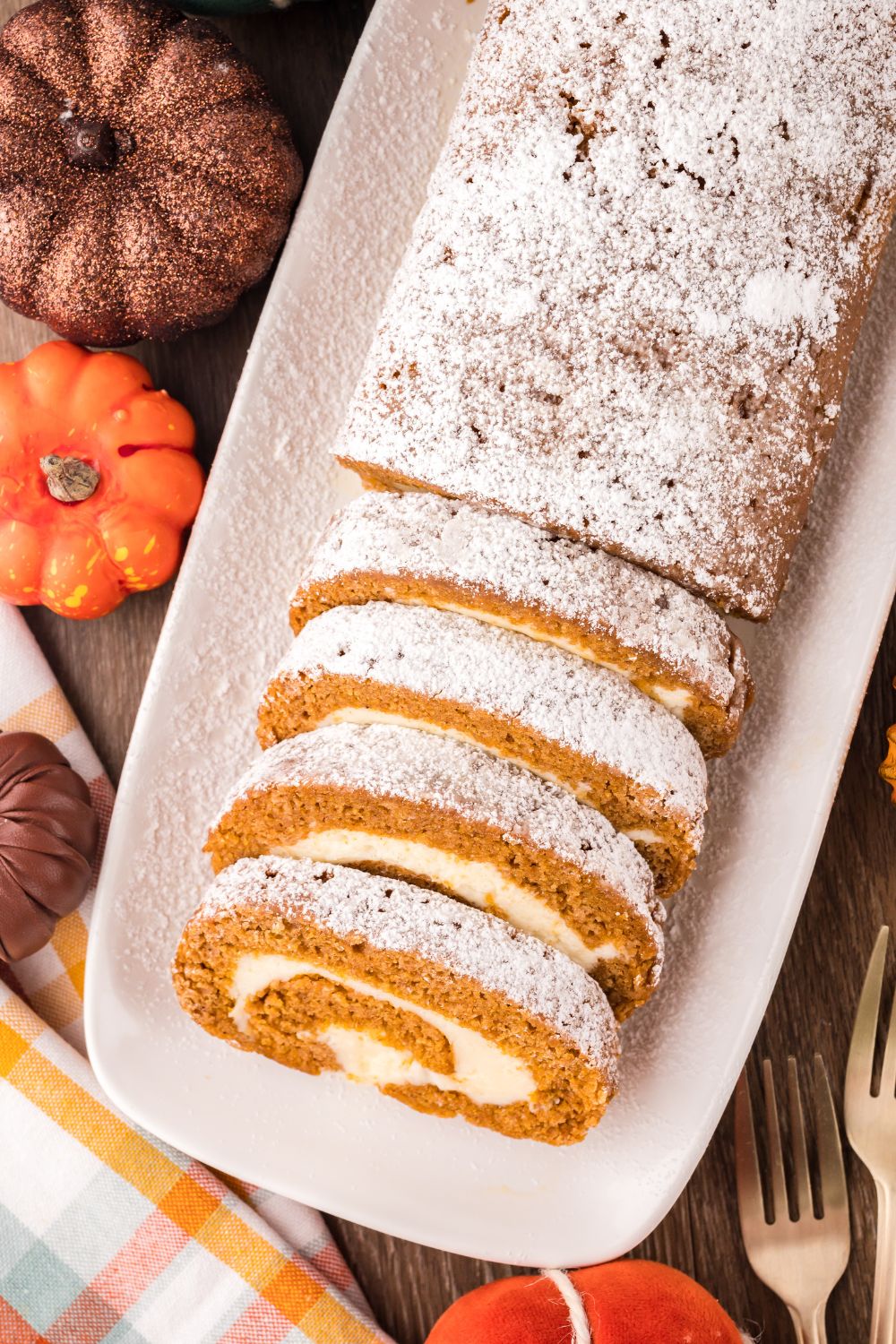 overhead look down on a wooden table with fake mini pumpkins and a long Pumpkin Cake Roll on a white platter. Part of the roll is sliced into wedges