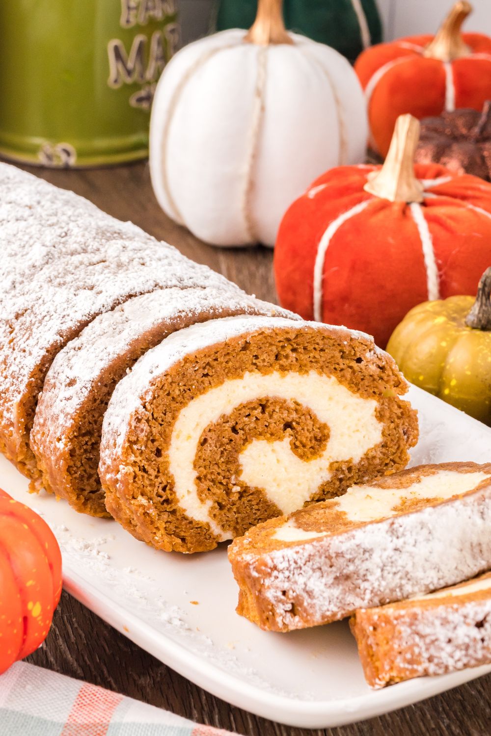 Pumpkin Cake Roll placed on table sitting on a white platter, with a portion of the pumpkin cake sliced. Then you have a mix of faux pumpkins scattered on table for a rustic fall look