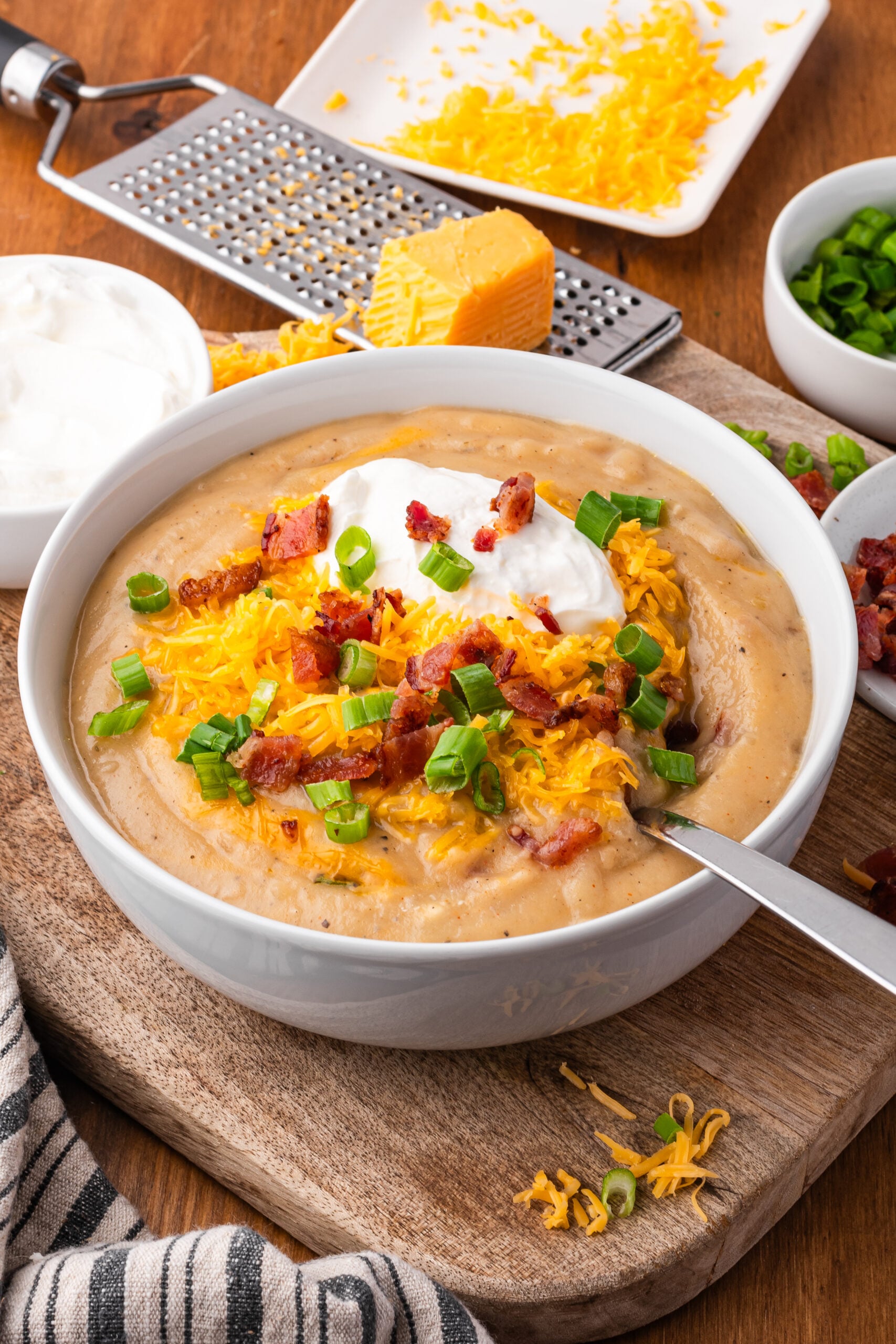 Crock pot loaded baked potato soup in a white bowl on a dark wood table with cheese grated in the background