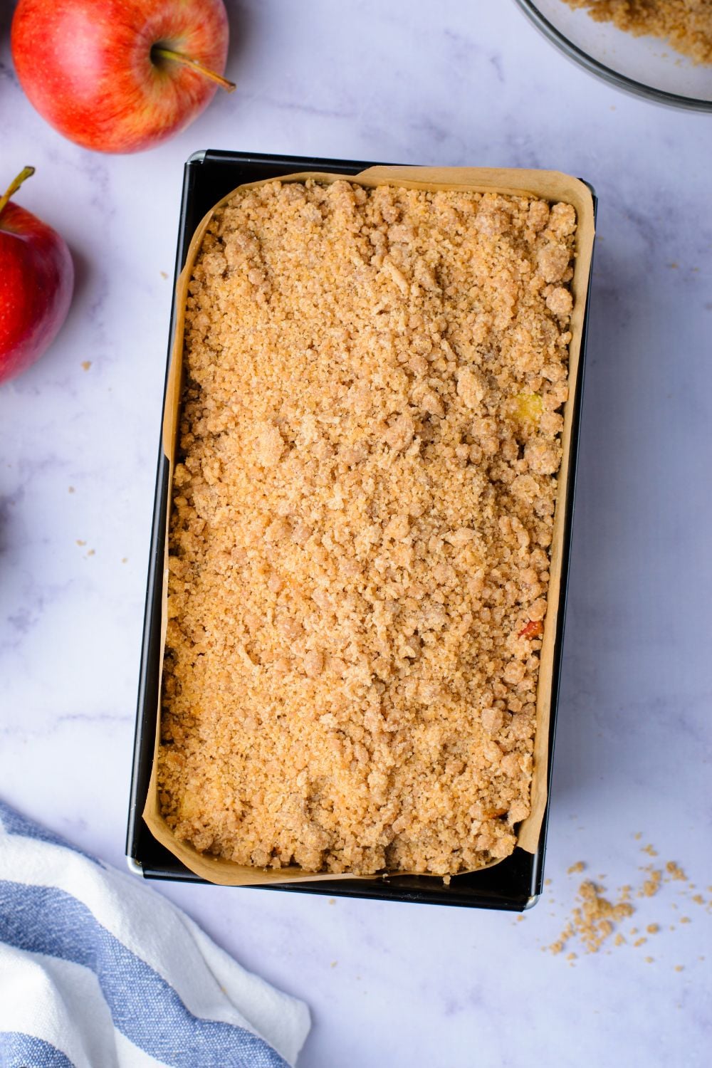 cinnamon crumble on top of a loaf cake batter in pan with a couple apples around the table