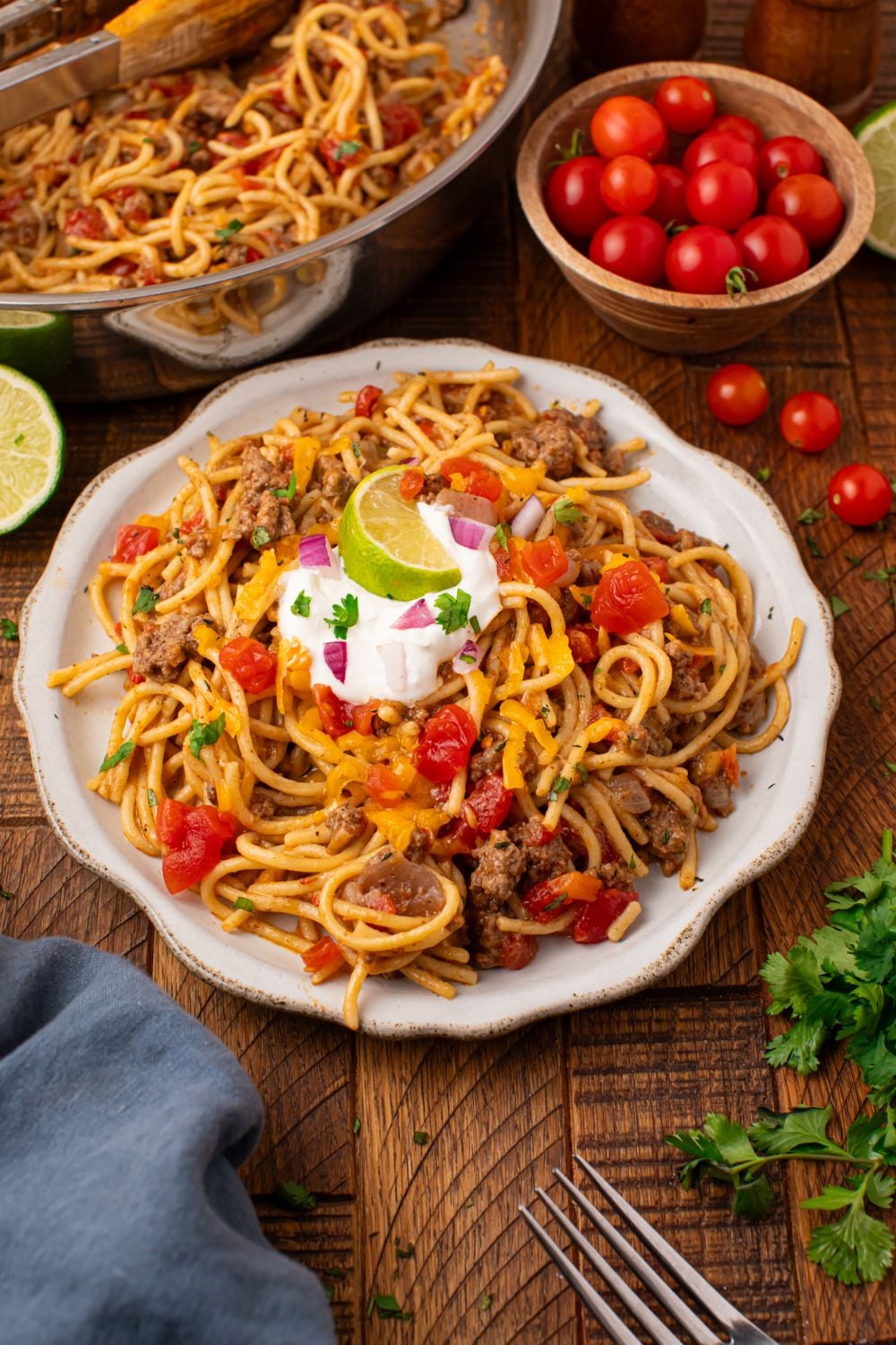 A plate with a pile of pasta with tomatoes, sour cream, a lime wedge on top, and more then a small tomatoes in a bowl with cilantro
