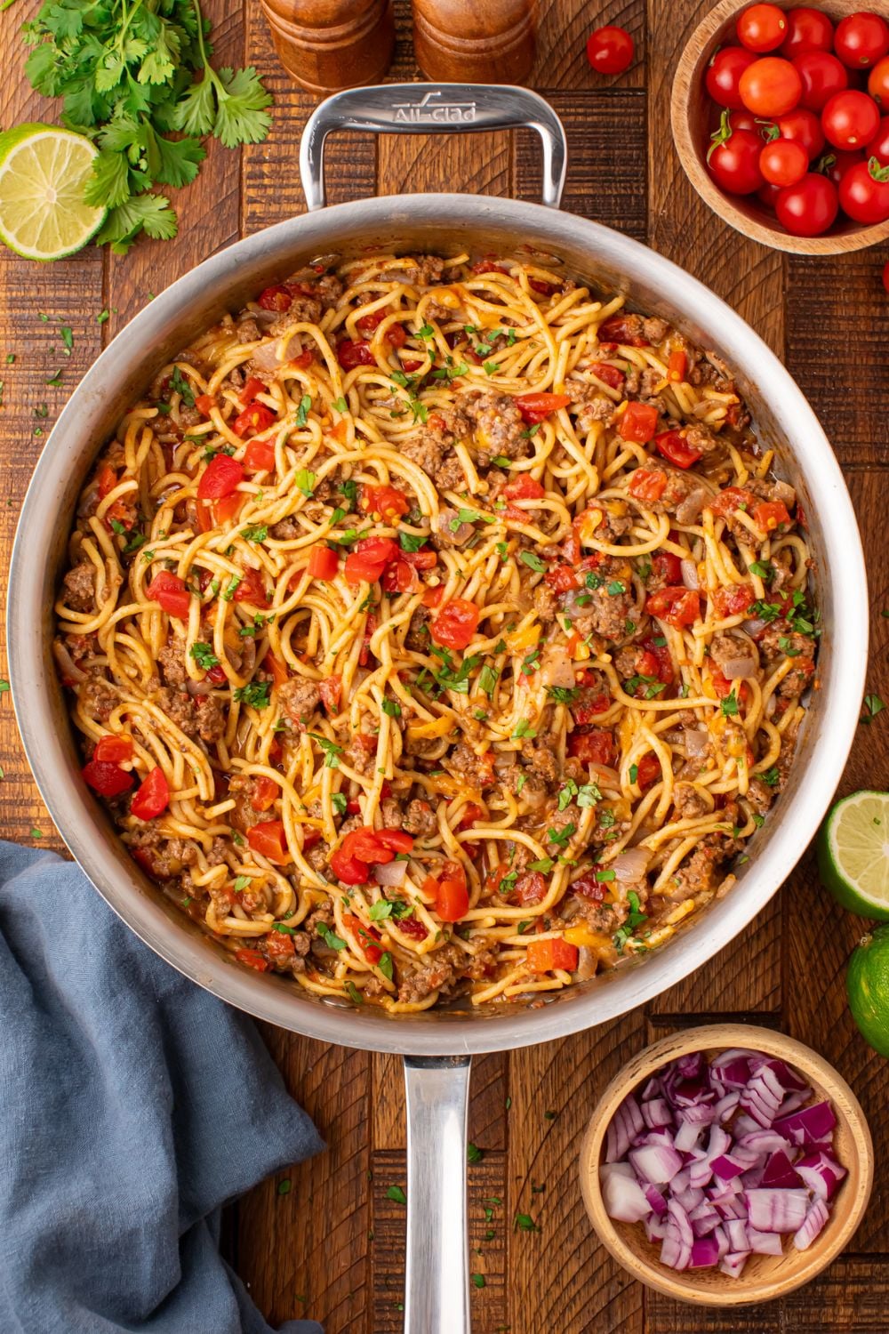 looking down on wooden table with a large metal skillet with taco spaghetti. Then cilantro, tomatoes, lime and more around table