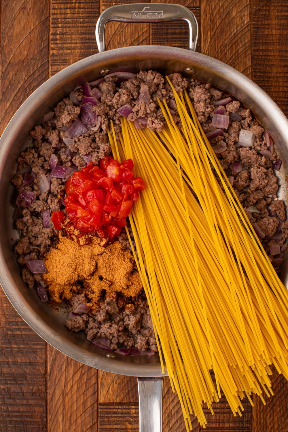 ground beef, tomatoes, taco seasoning and dry pasta in a skillet on wooden table
