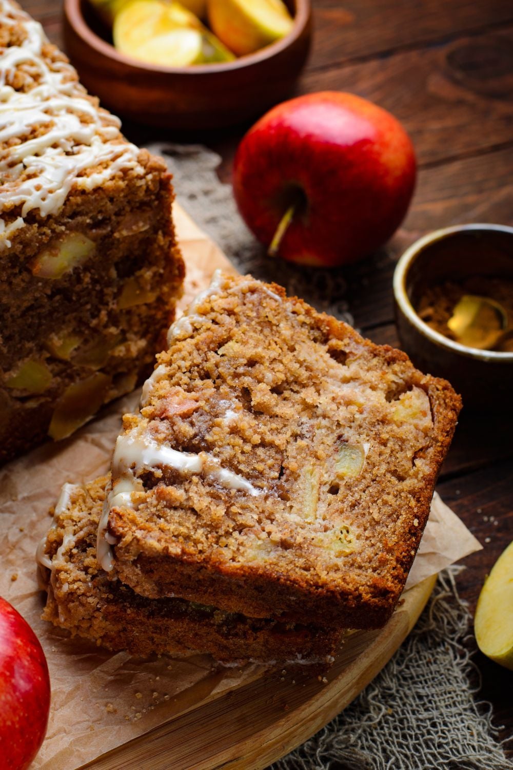 Apple Cinnamon Loaf Cake on a wooden table and some of the slices are stacked on top of one another with apples sliced in a wood bowl and a whole apple to side