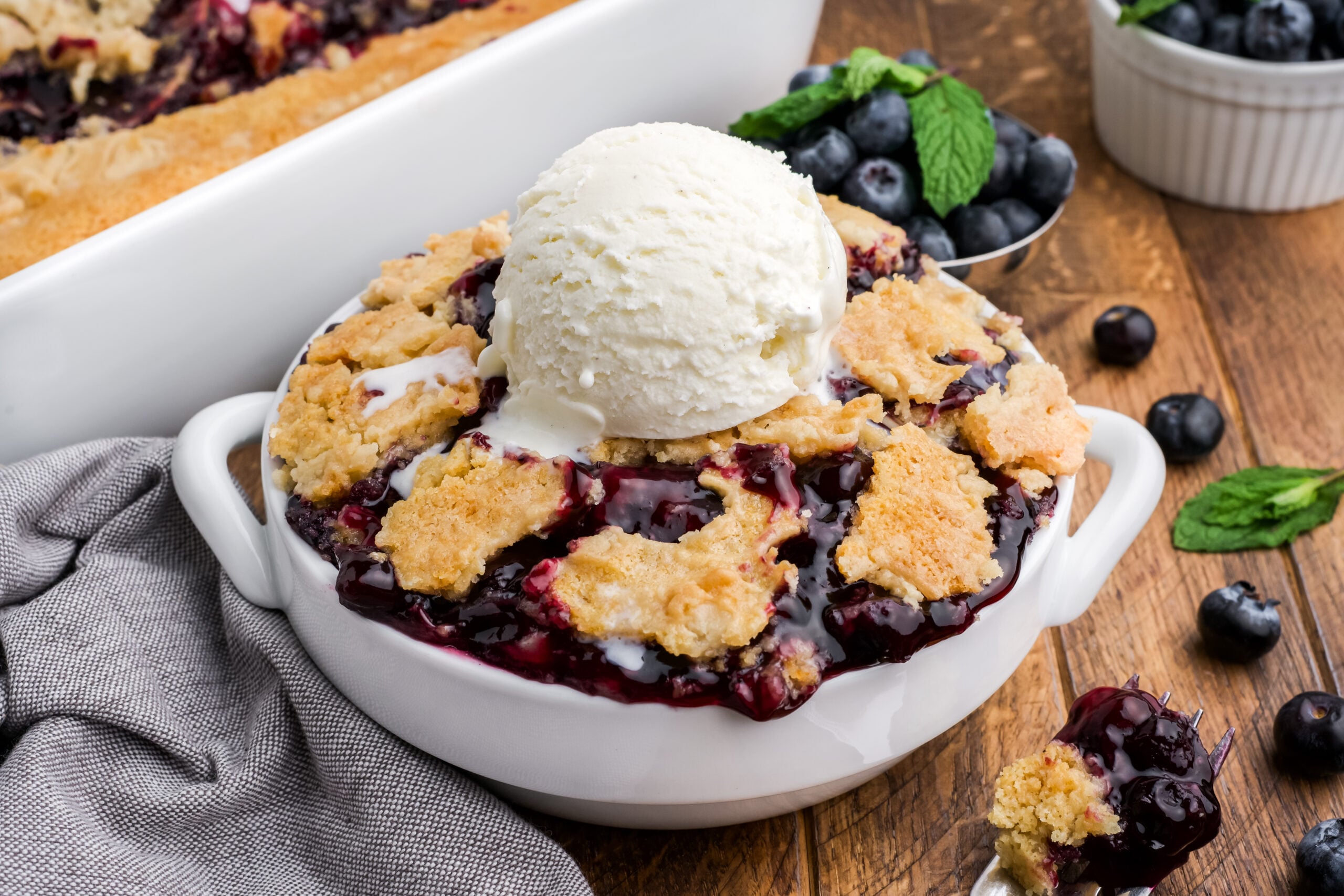 looking down on wooden table with a bowl of Blueberry Dump Cake with a scoop of ice cream in center