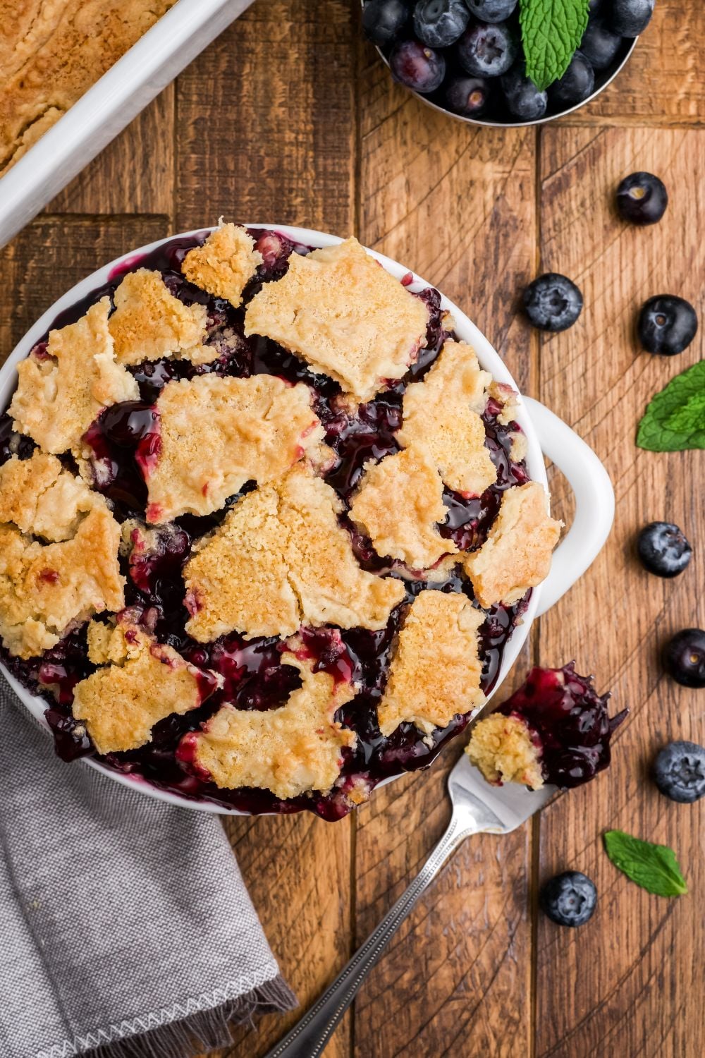 white bowl overflowing with blueberry dump cake and a fork with more cake with more blueberries scattered on table