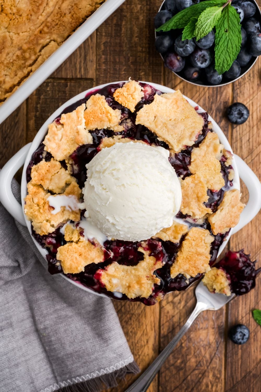 looking down on wooden table with a bowl full of dump blueberry cake with ice cream in the center and a bowl of blueberries to side