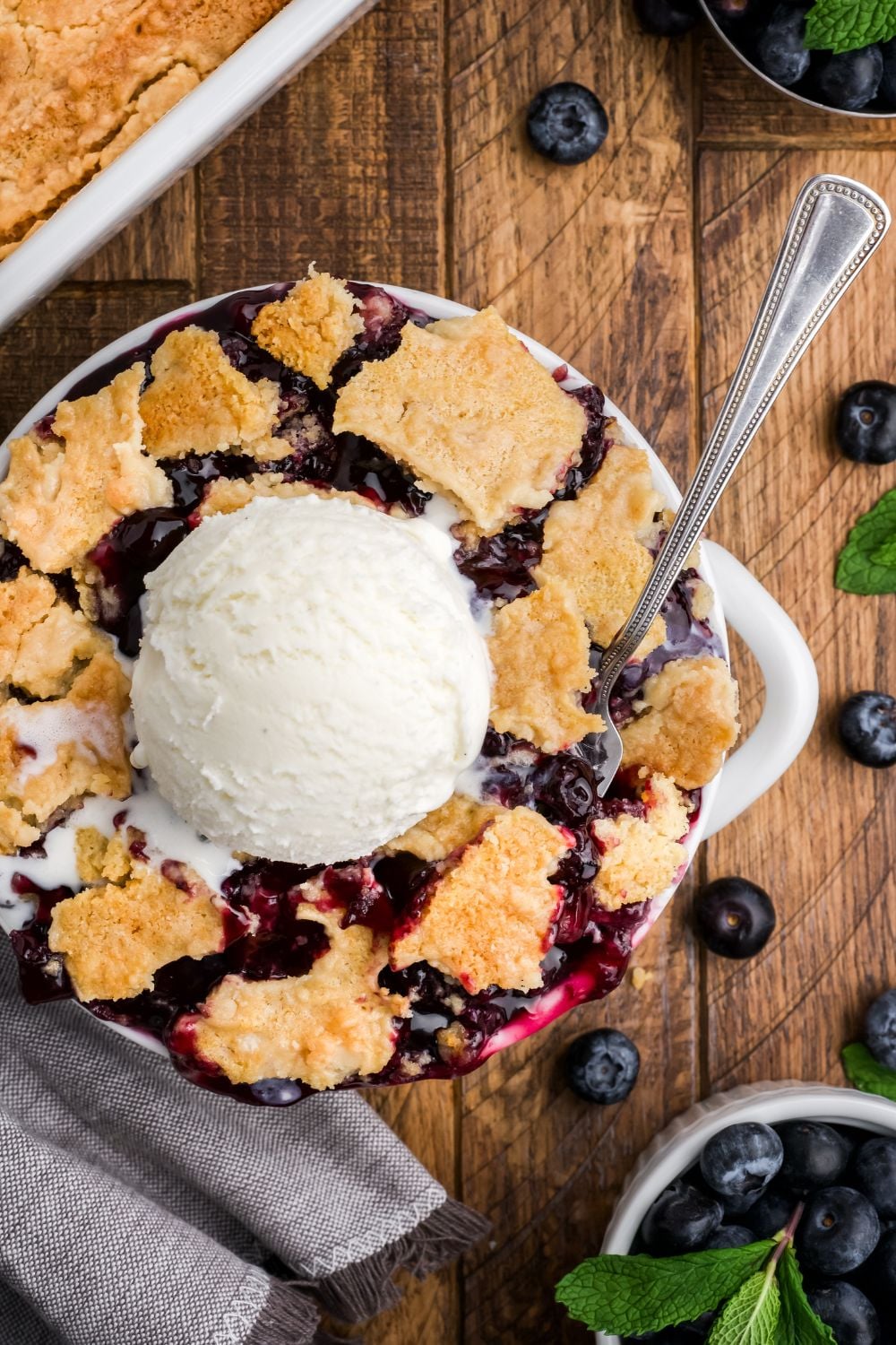 looking down on wooden table with a bowl of Blueberry Dump Cake with a scoop of ice cream in center