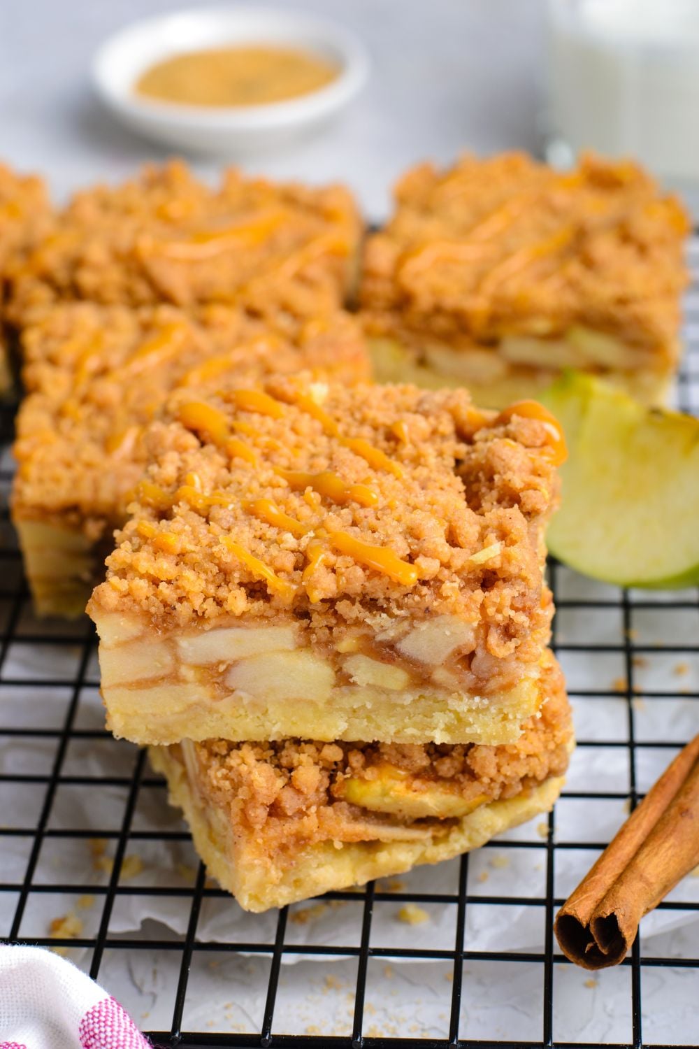 Caramel Apple Pie Bars stacked on top of one another and more bars around the cooling rack with cinnamon sticks and sliced apples