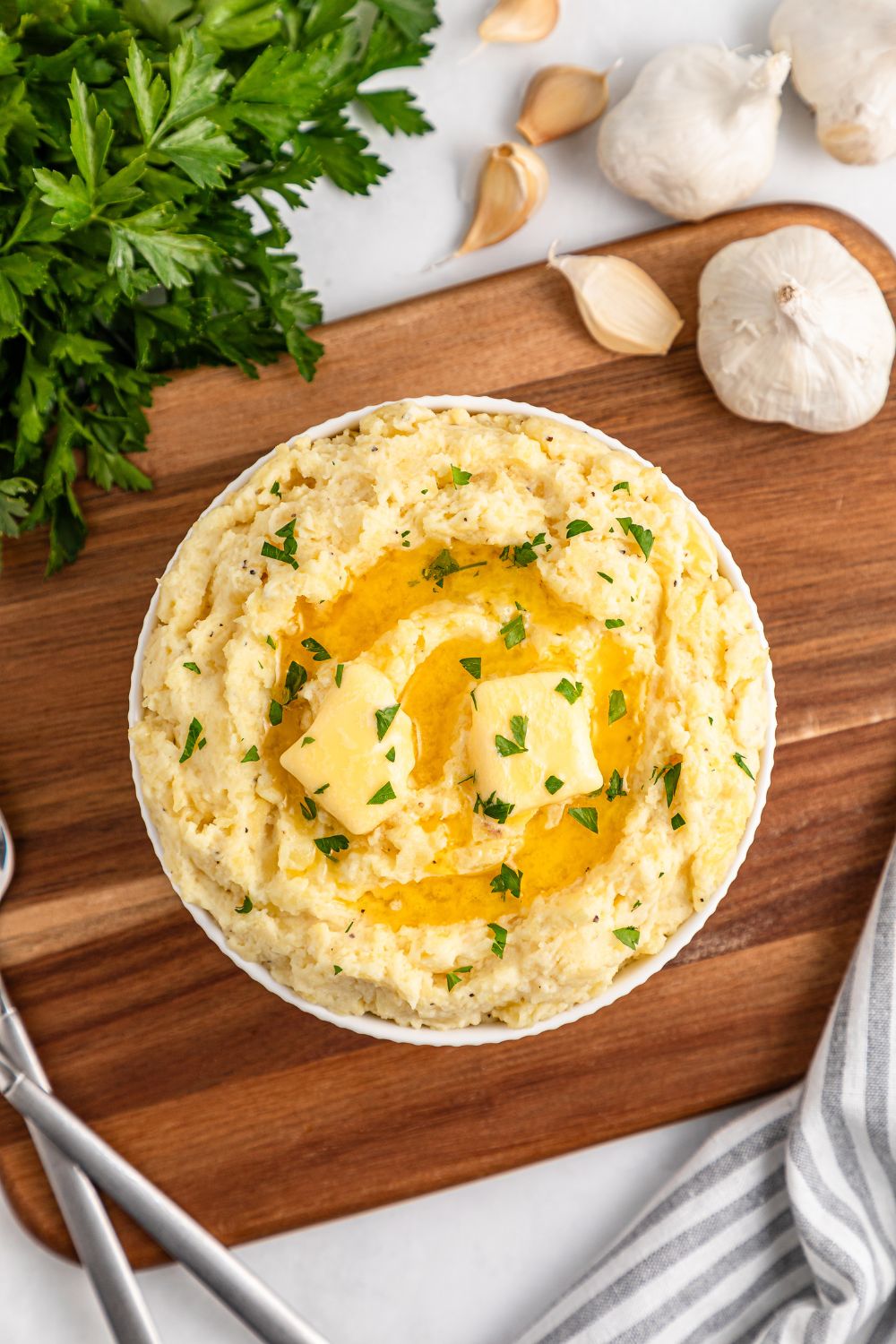 Looking down at table to see a wooden cutting board with a bowl of Garlic Mashed Potatoes in the center. To the side a bundle of parsley and fresh garlic cloves