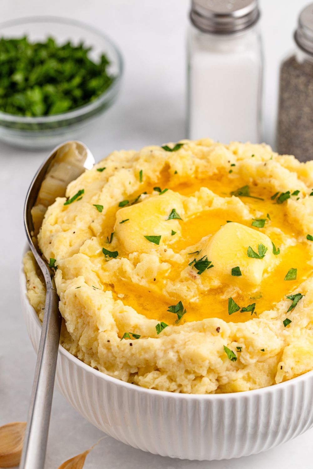 a white bowl sitting on table full of thick and creamy Garlic Mashed Potatoes with a spoon on its side wedged into the bowl