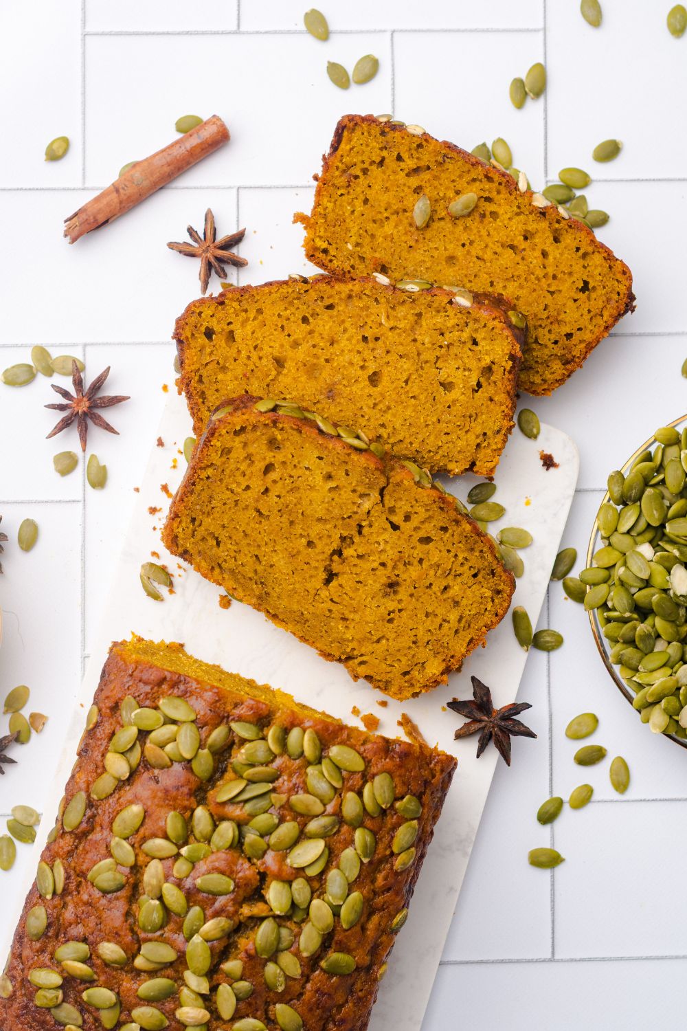looking down on table to a loaf of Starbucks Pumpkin Bread with three slices placed on table with pumpkin seeds, and cinnamon sticks around the table