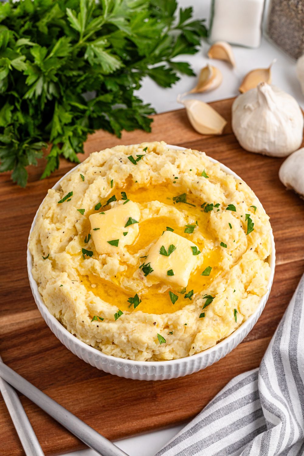 a wooden cutting board with a white bowl full of Garlic Mashed Potatoes in it, topped with melted butter and parsley. To the side is fresh garlic, a towel, and parsley
