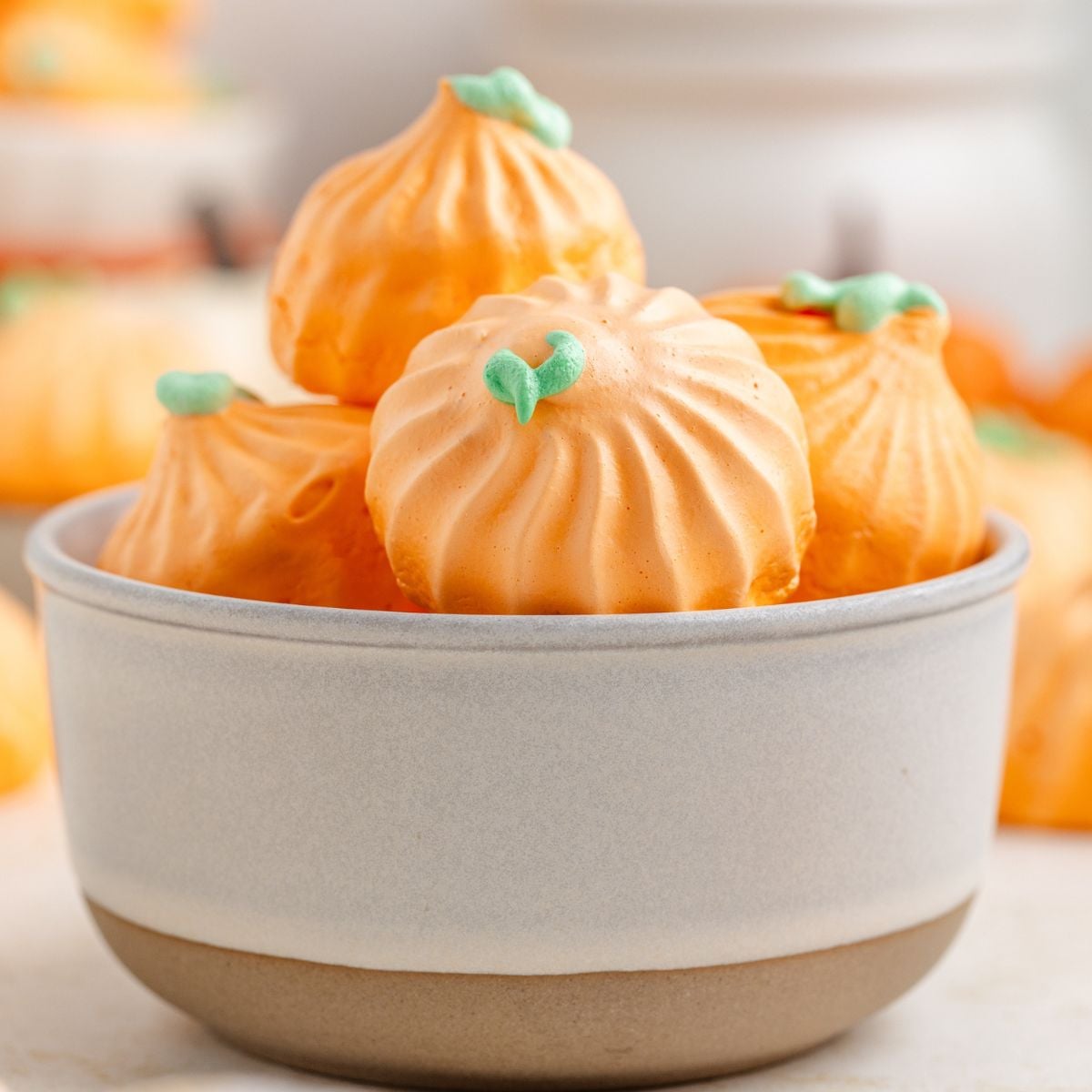 Pumpkin Meringues placed in a white bowl on table with more meringues behind it on table 
