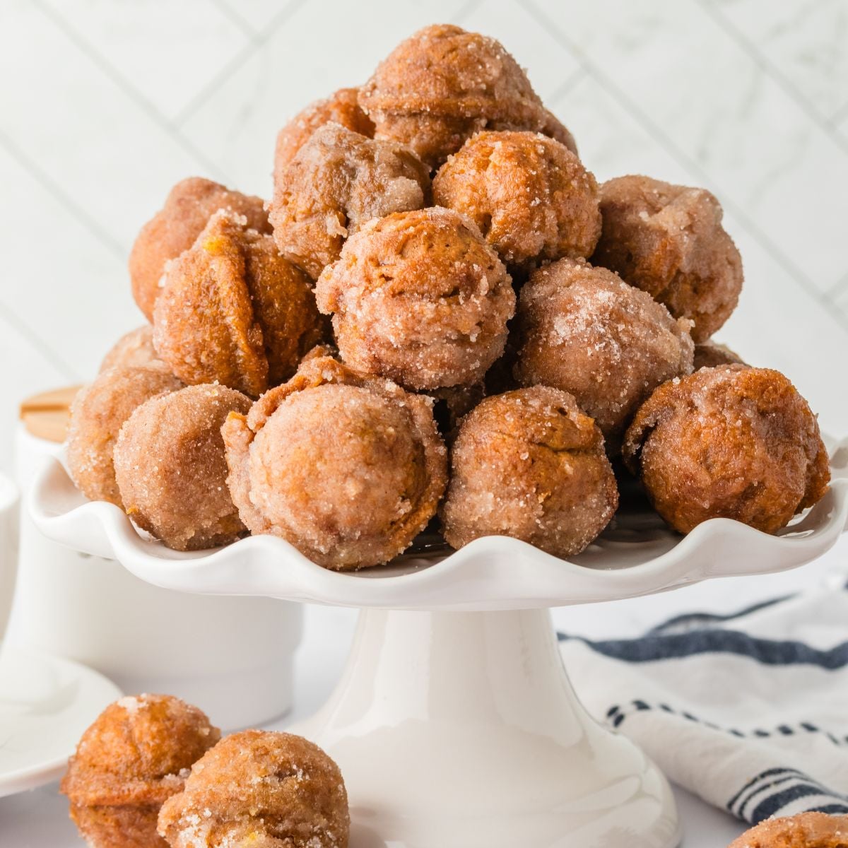 Pumpkin Spice Donut Holes stacked on a cake platter sitting on table with more donut holes scattered around the table 