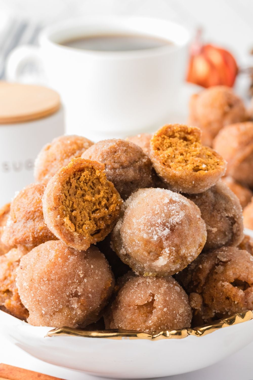 Pumpkin Spice Donut Holes stacked in a bowl on the counter with a few cut in half, and a cup of coffee in the background of the image 