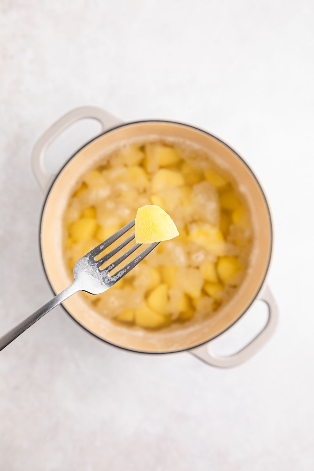 fork holding a potato above a pot full of potatoes and water on table