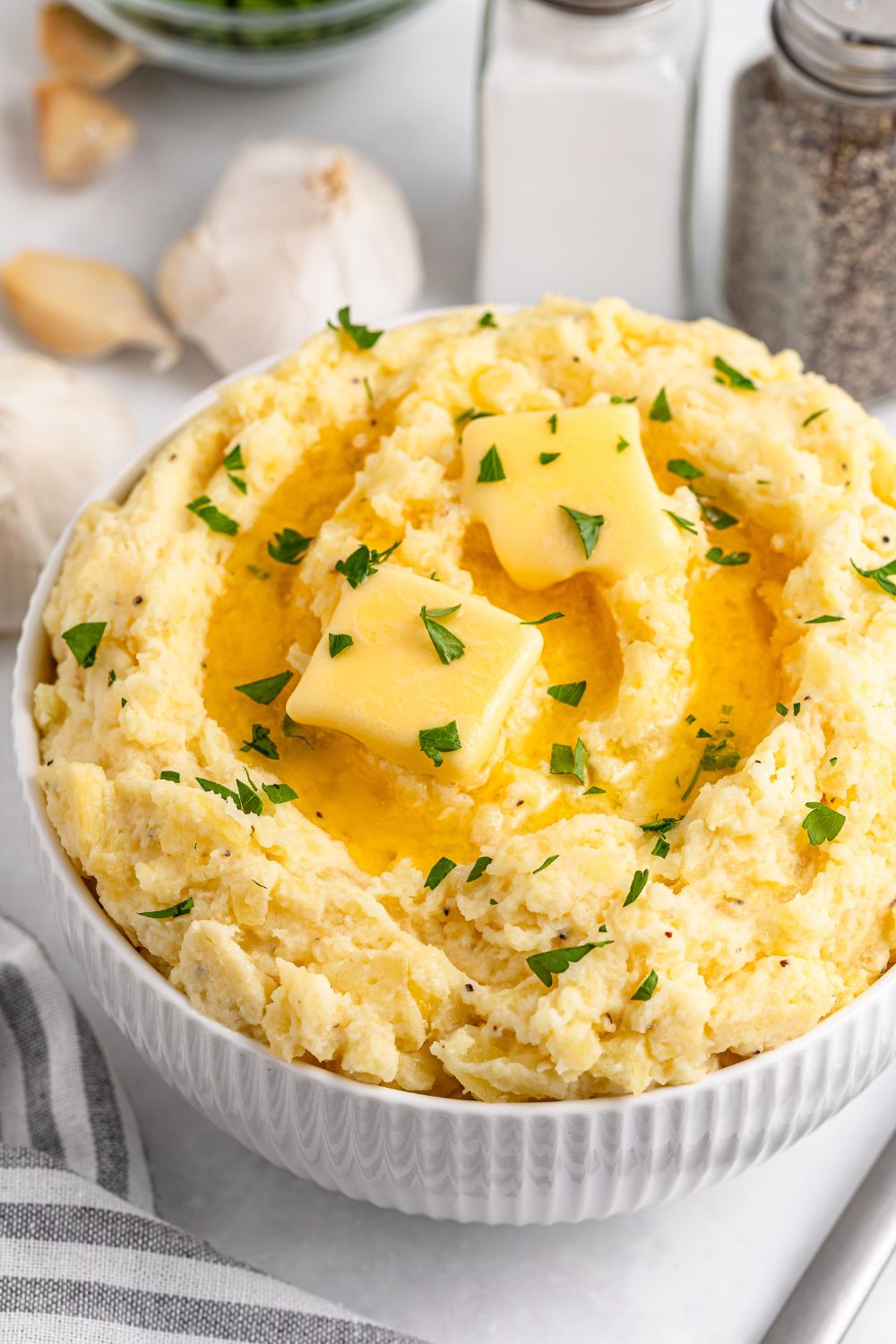 Garlic Mashed Potatoes in a bowl on white table with garlic and a striped towel to side of the bowl