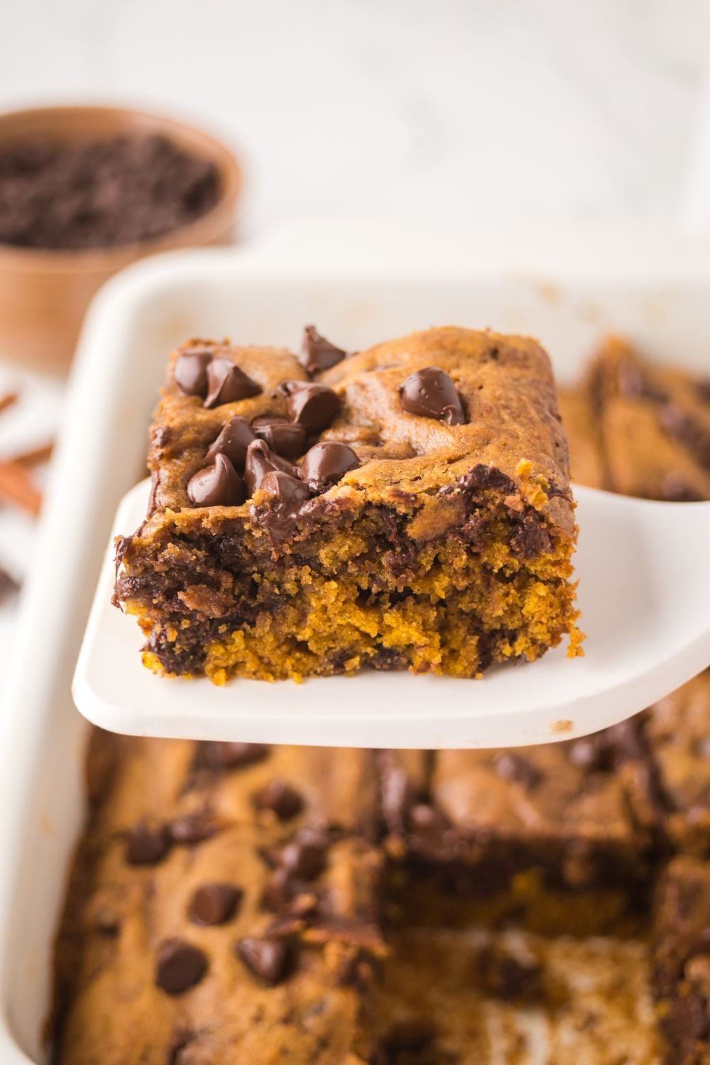 white spatula with a slice of Brown Butter Pumpkin Chocolate Chip Bar on it, and a pan full of bars sitting on table