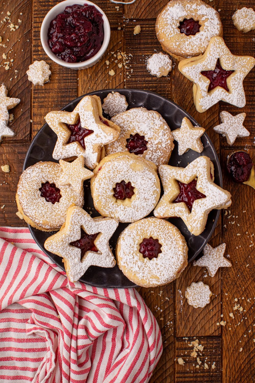 Linzer cookies scattered around on a wooden table with a bowl of jam to side and a red and white towel