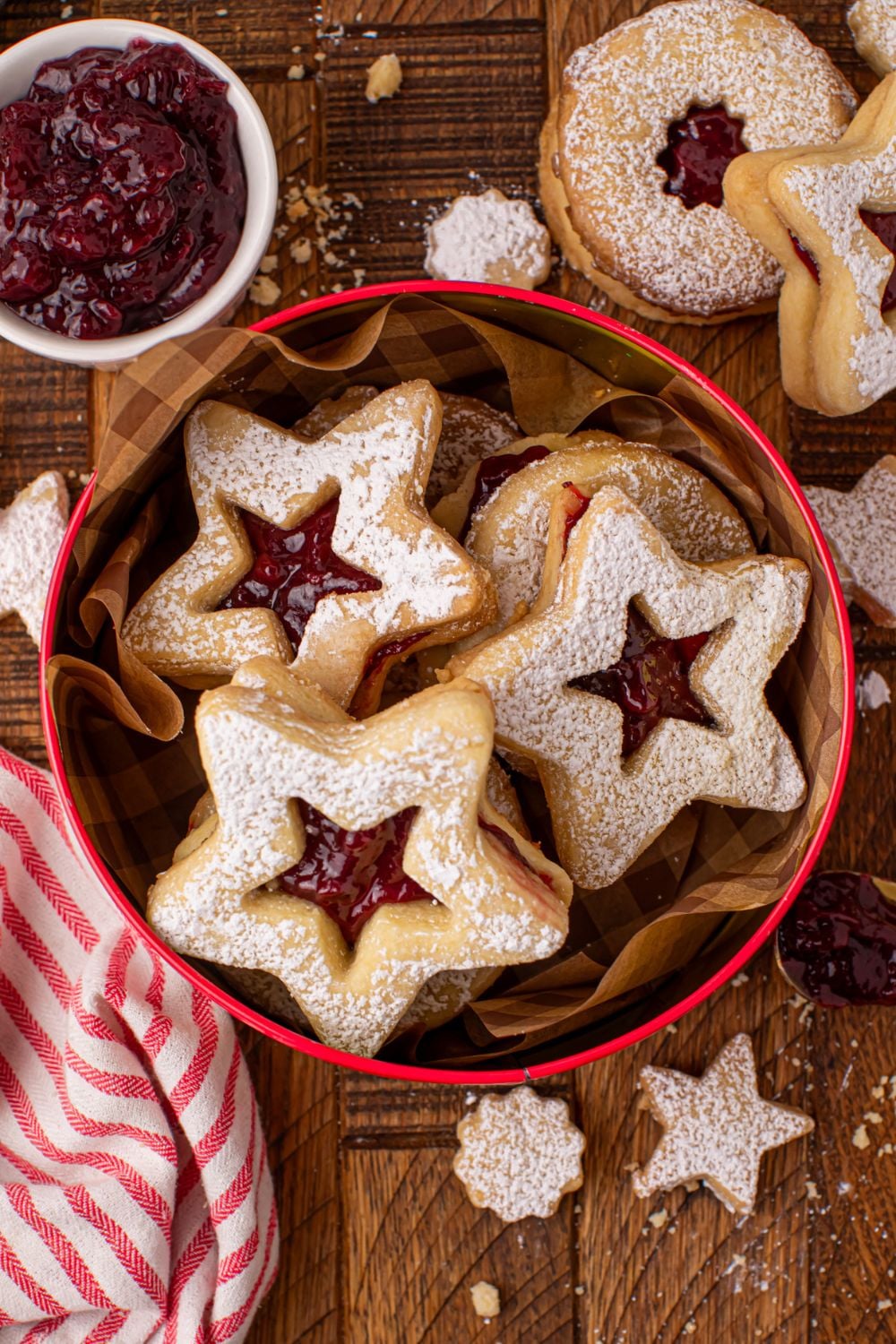 tin full of homemade Linzer cookies on a wooden table with more cookies scattered around table and a bowl of jam to side