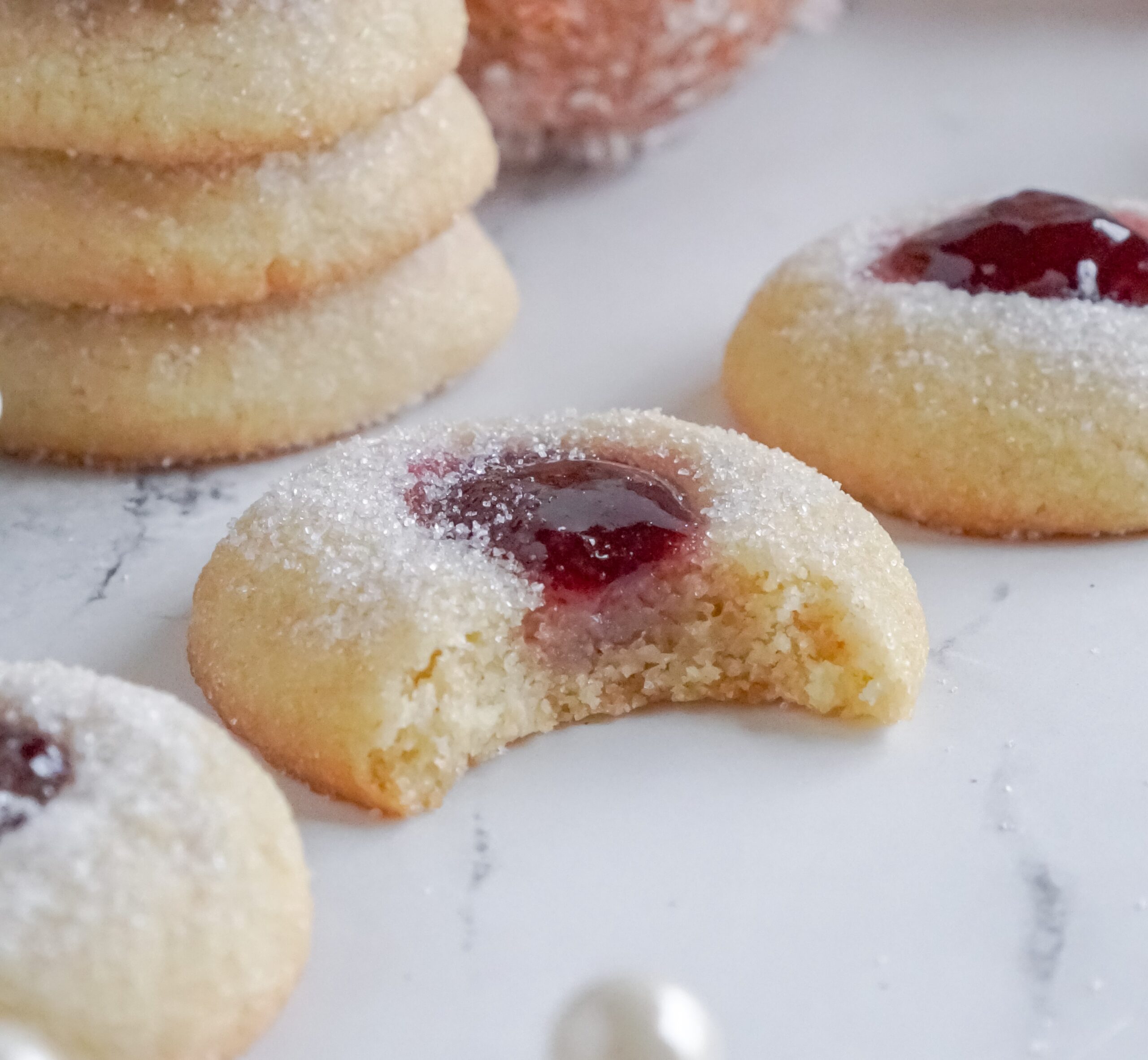 Raspberry Thumbprint Cookie with a bite missing on a marble table with more cookies spread around it