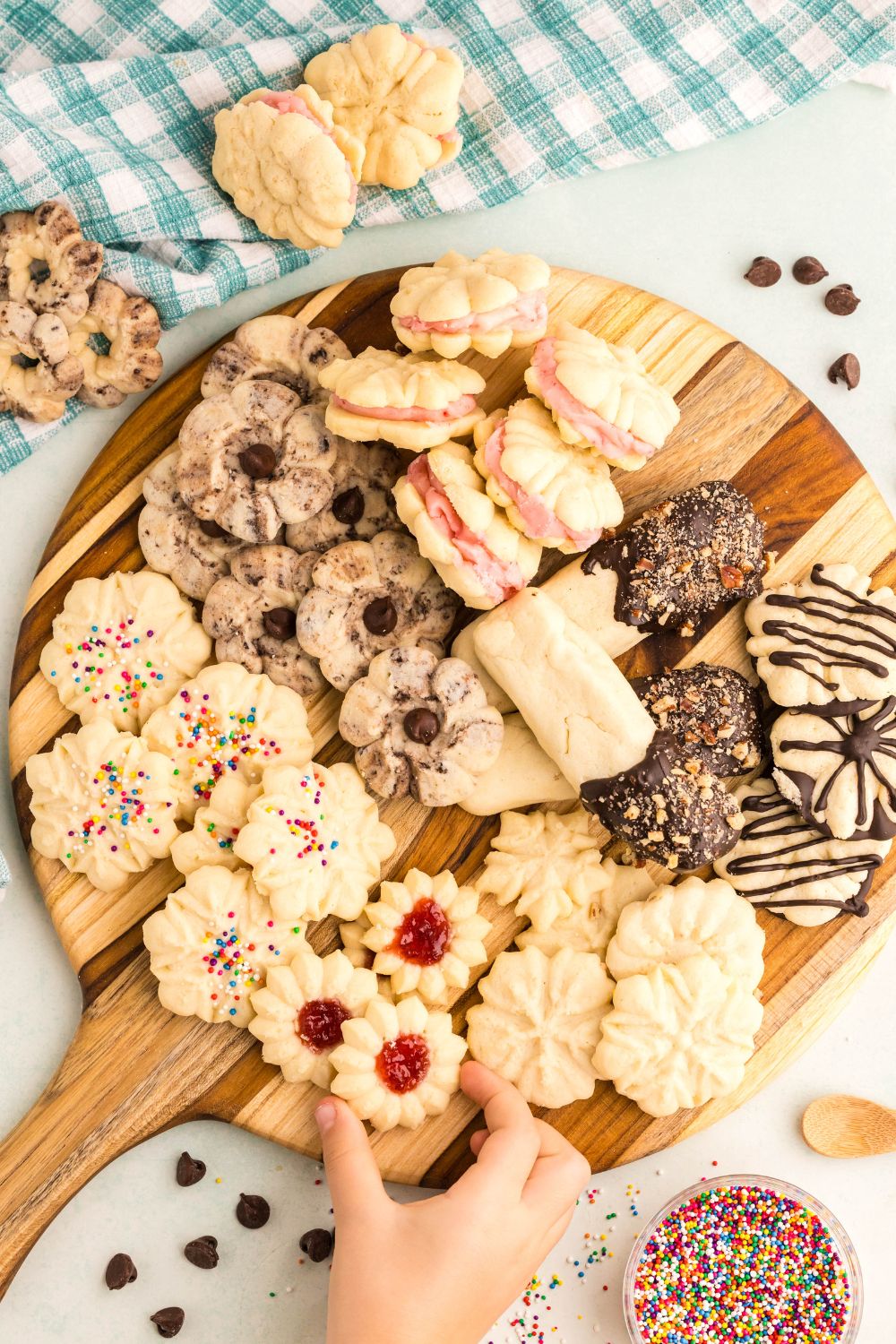 Spritz Christmas Cookies on a wooden serving tray on a white table with a towel behind it on table and a little kid grabbing a cookie