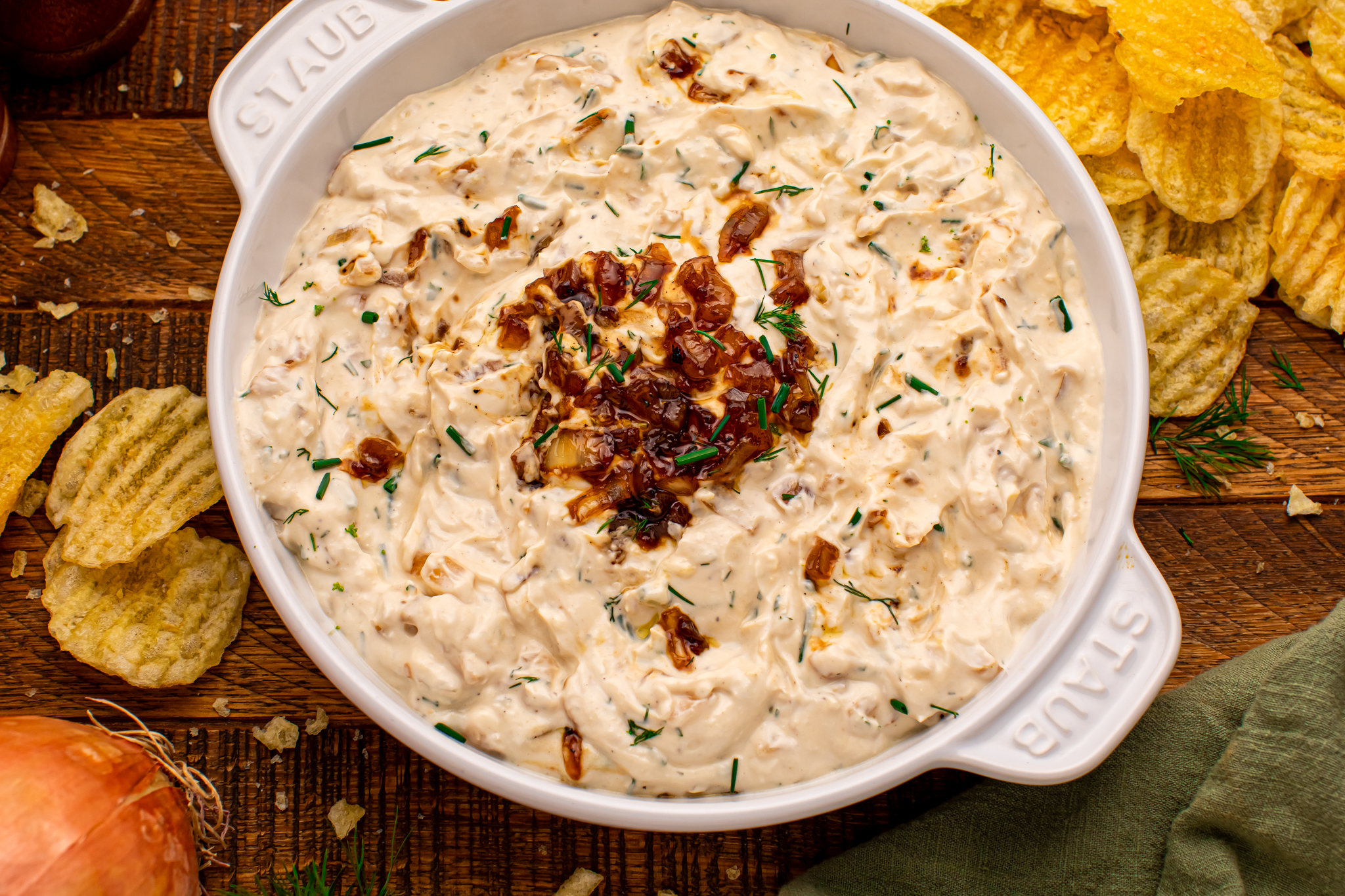 Overhead photo of Caramelized Onion Chip Dip in a white staub bowl on dark wooden table with chips all around the bowl