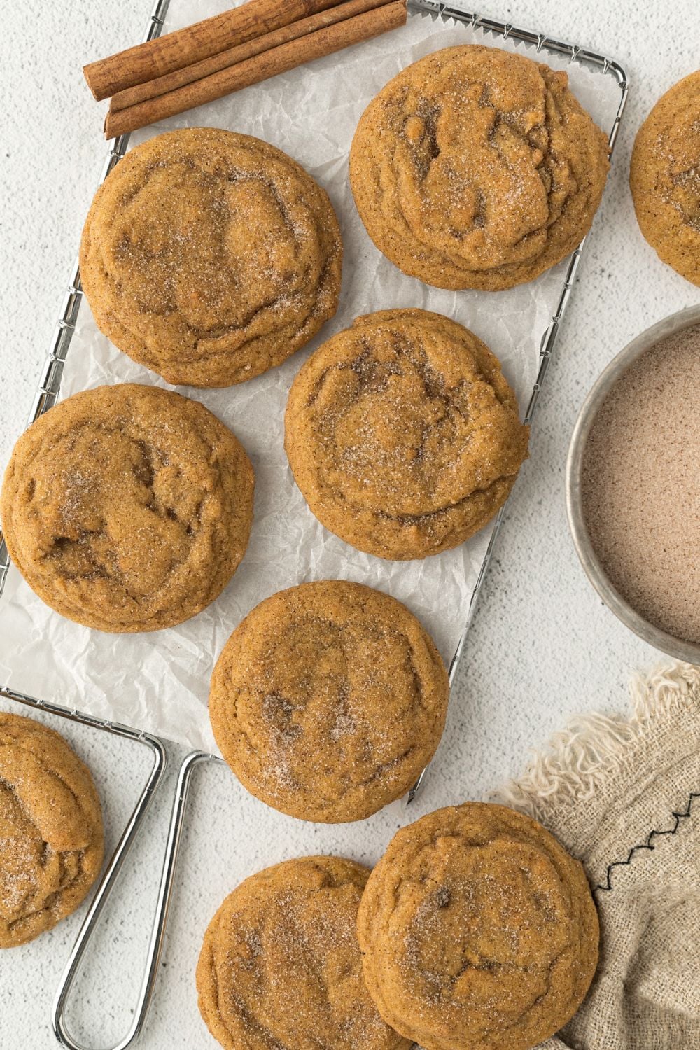 Pumpkin Snickerdoodle Cookies spread out on a white table with cinnamon sugar to side and cinnamon sticks around cookies 