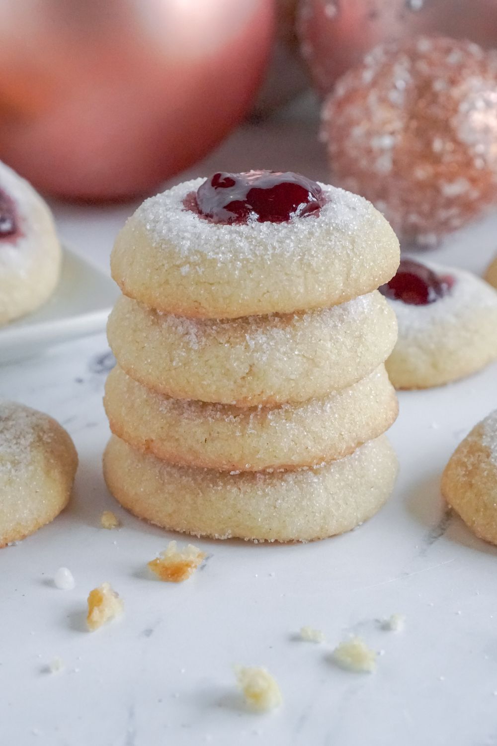 Raspberry Thumbprint Cookies stacked on top of another with more cookies spread around table with crumbs scattered and Christmas balls