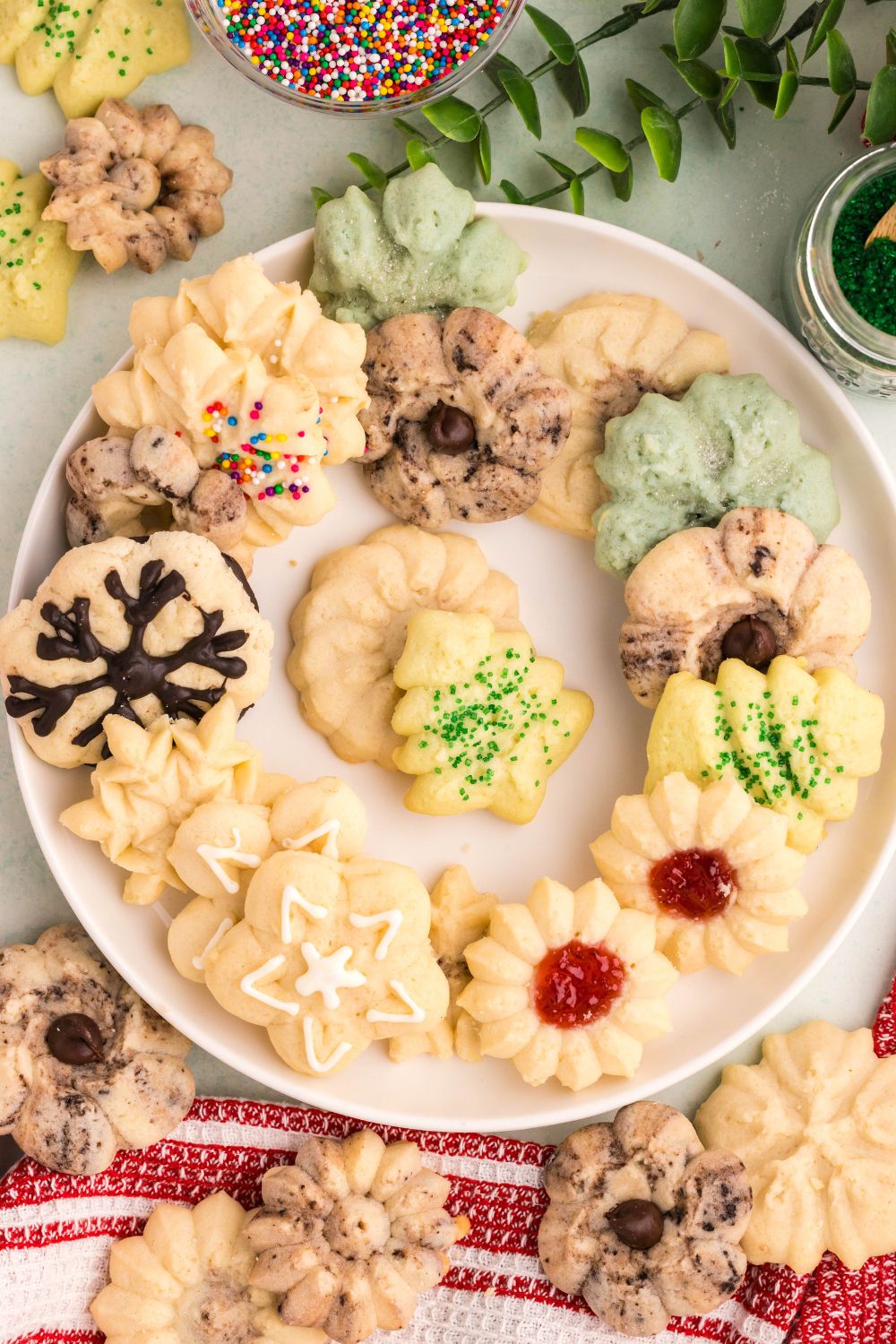 a white plate full of a mix of decorated Spritz Christmas Cookies with more cookies around the table with a towel under some of the cookies