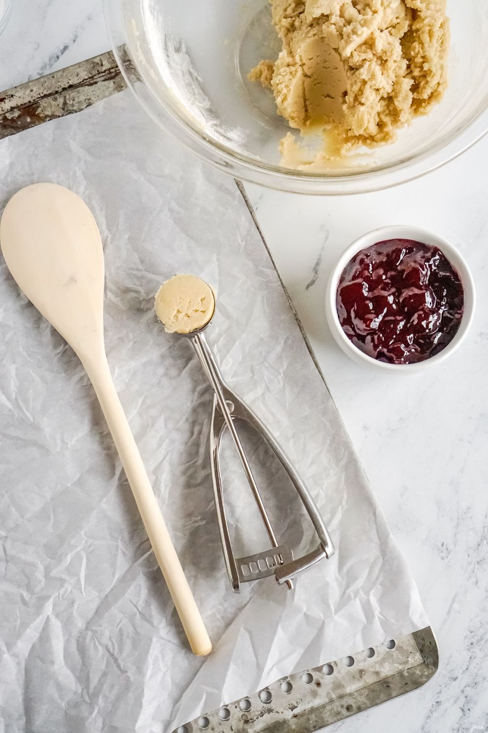 Raspberry Thumbprint Cookie dough in a glass bowl, with jam in a small bowl to side and cookie baking sheet on table