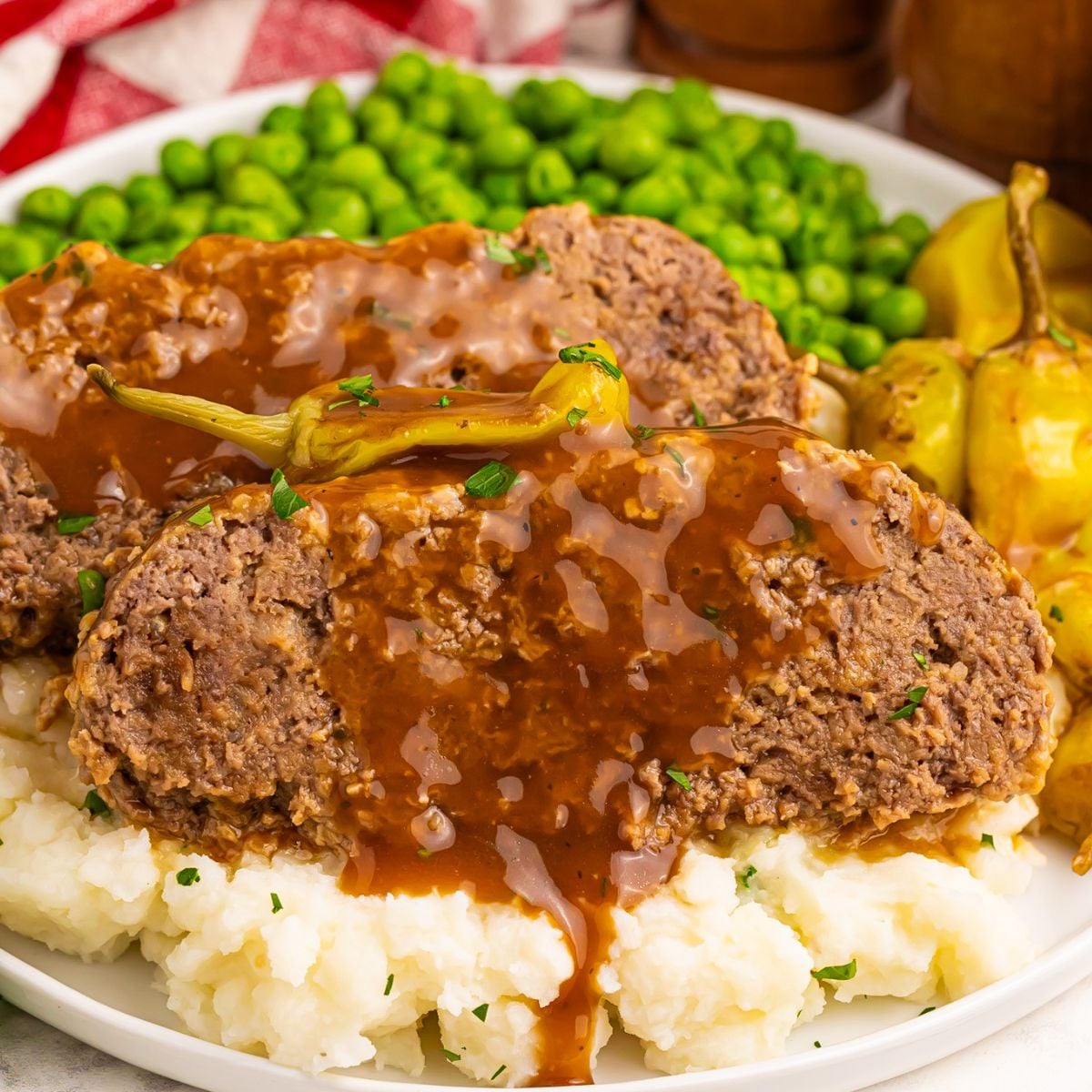 Mashed potatoes topped with Crockpot Mississippi Meatloaf and a side of green peas on a large plate on a wooden table
