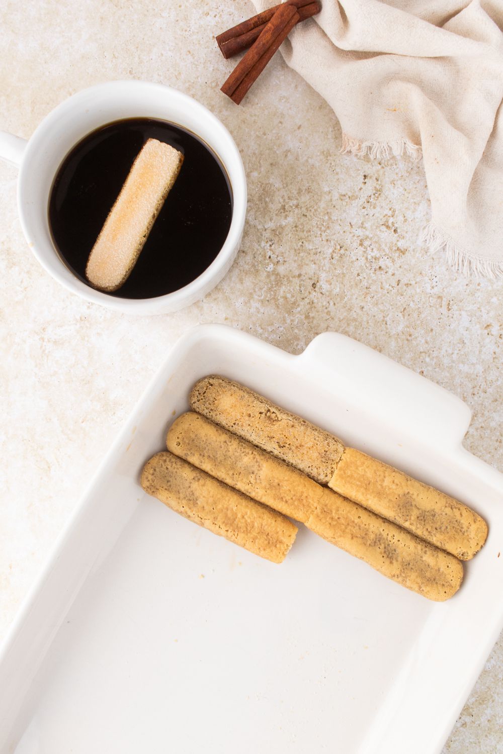 a bowl of coffee to side for dipping ladyfinger cookies with a casserole dish with a layer of cookies being placed in it 