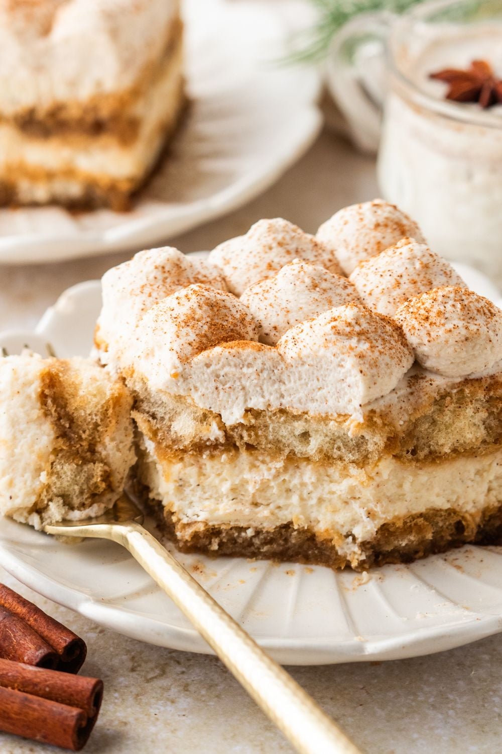 Eggnog Tiramisu sliced into a square on plate wit ha bite on a fork on side of plate with cinnamon sticks on table 
