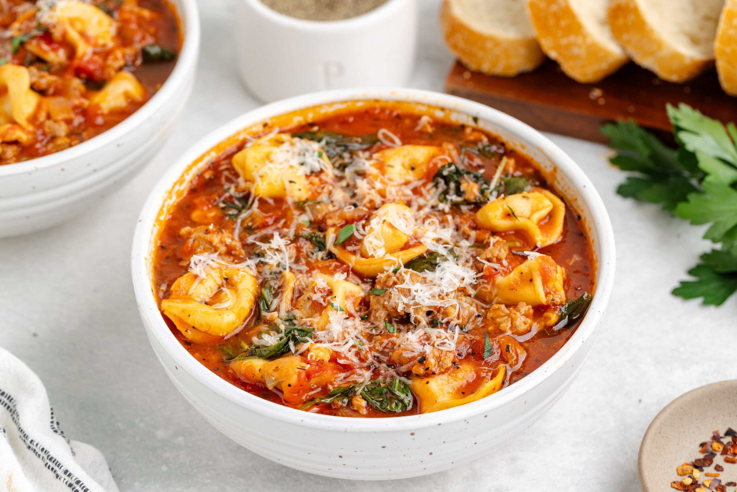 Sausage Tortellini Soup in a white bowl topped with fresh grated parmesan cheese on a white marble table