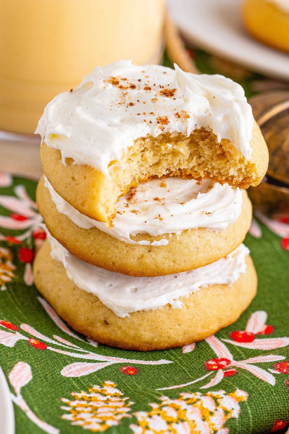 Soft Frosted Eggnog Cookies stacked on table with a bite taken out of it and a glass of eggnog to side of table