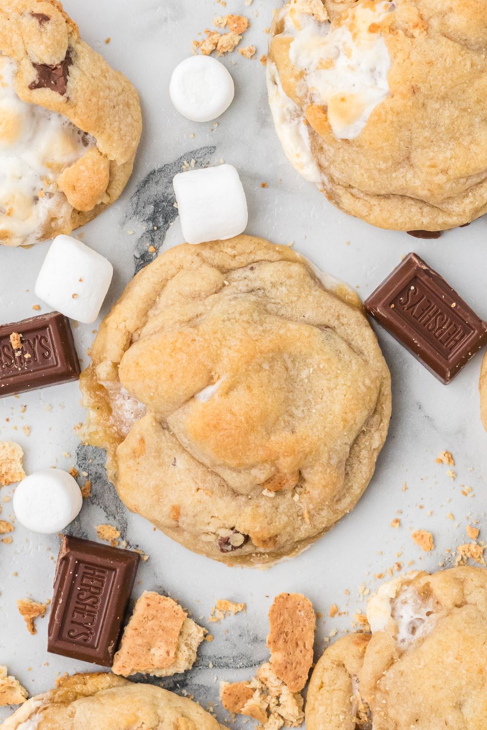bakery style s'mores cookies spread around the marble counter with graham crackers, marshmallows, and chocolate around table
